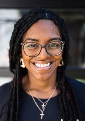 Close-up of a smiling woman with glasses, dreadlocks, and jewelry.