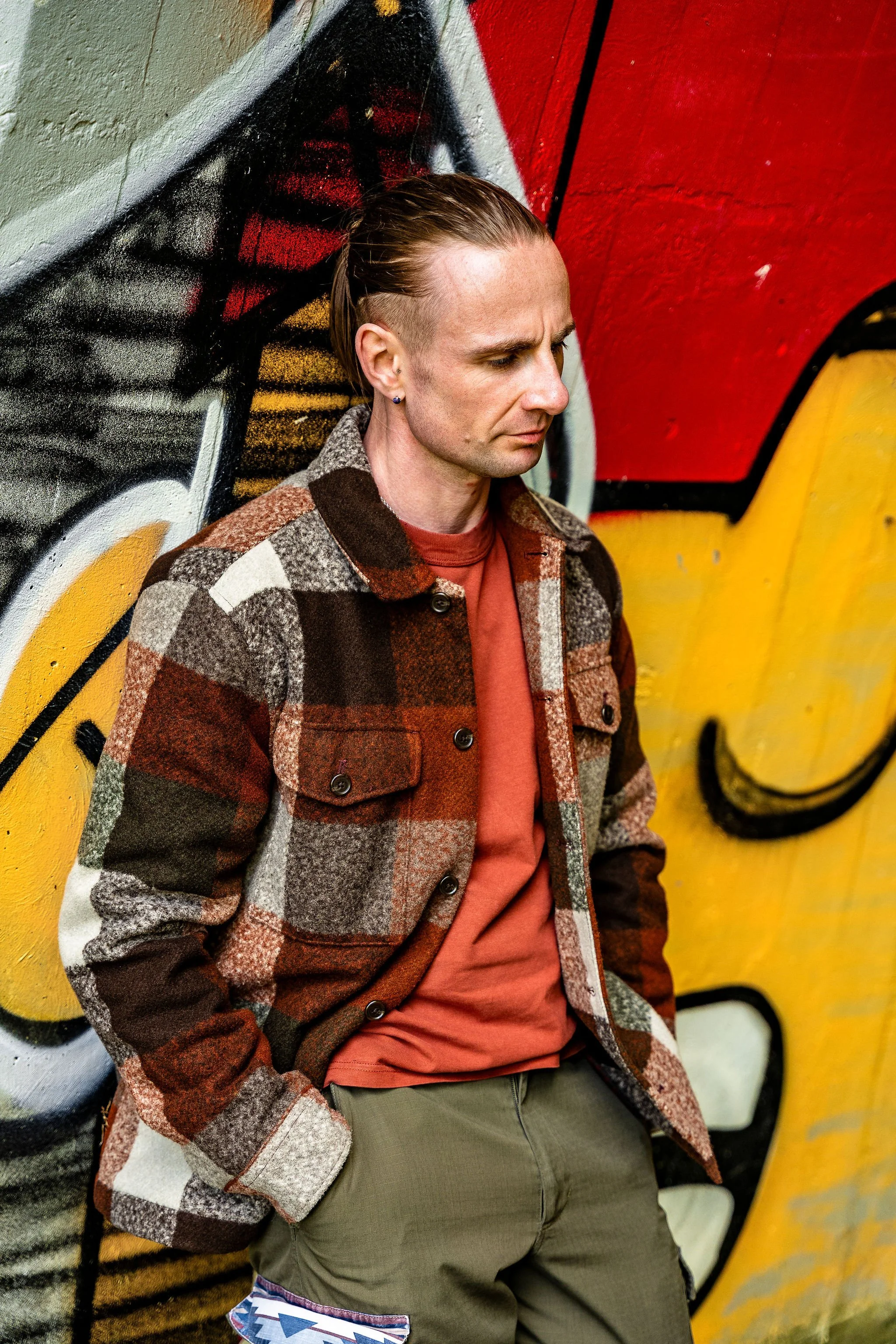 A man with slicked-back hair, wearing a plaid jacket over a red shirt, standing in front of a colorful graffiti wall.