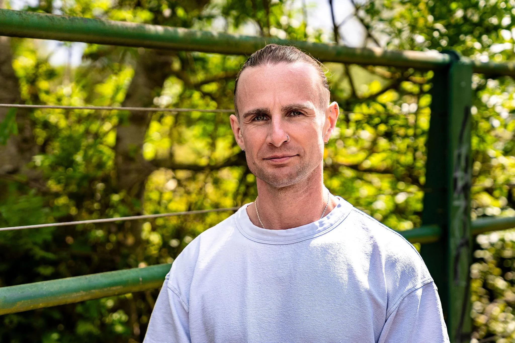A person with a buzz haircut and earrings standing outdoors in front of green trees and a metal railing, wearing a white t-shirt.