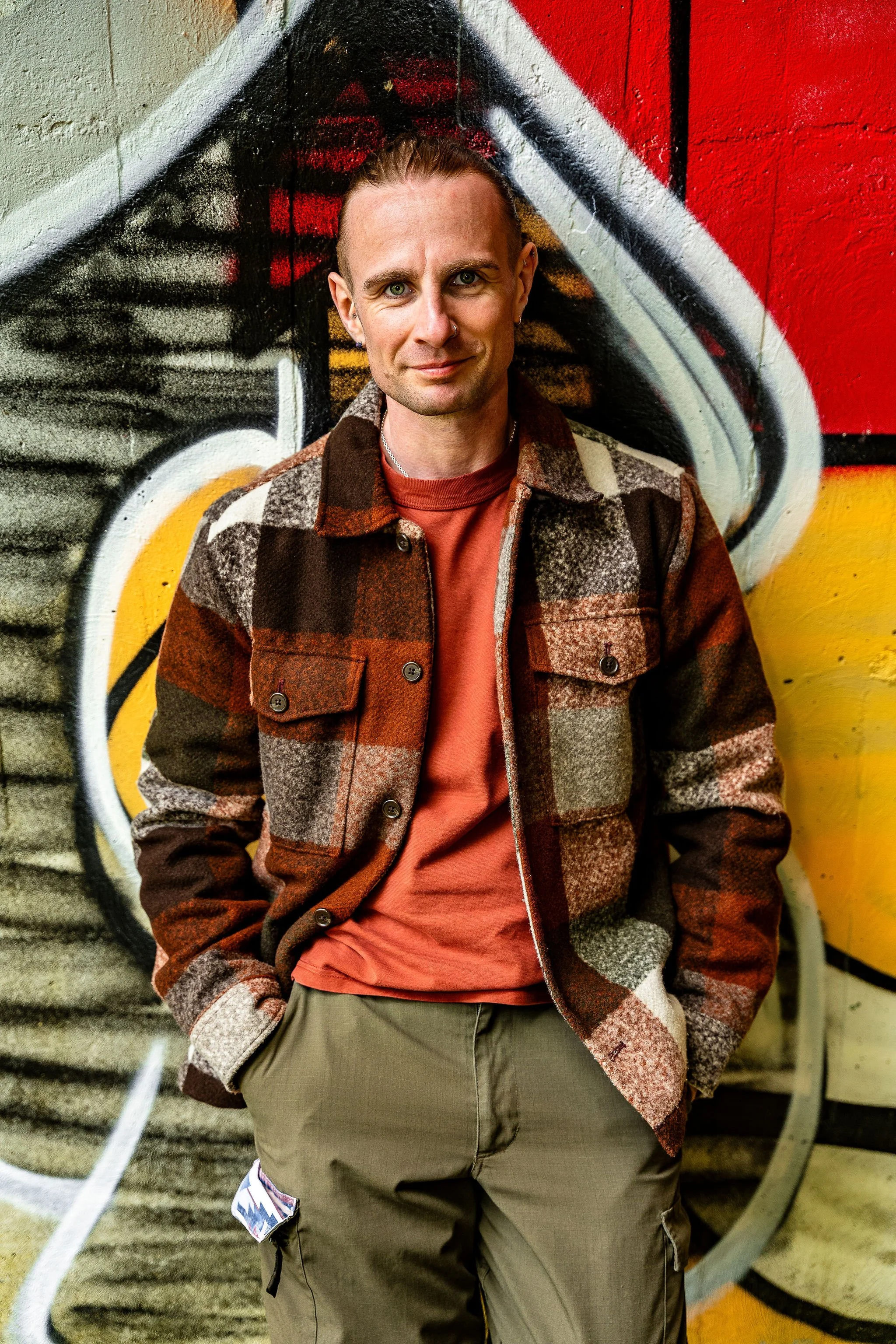 A man with short hair and earrings wearing a plaid jacket over a red shirt, standing in front of a colorful graffiti wall.