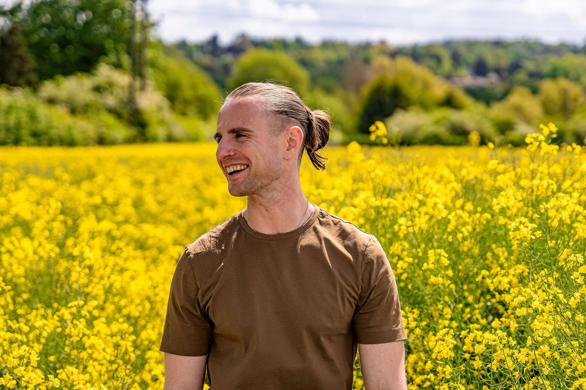 A young man with a man bun, wearing a brown t-shirt, smiling and looking to the side, standing in a field of yellow flowers with a backdrop of green trees and hills.