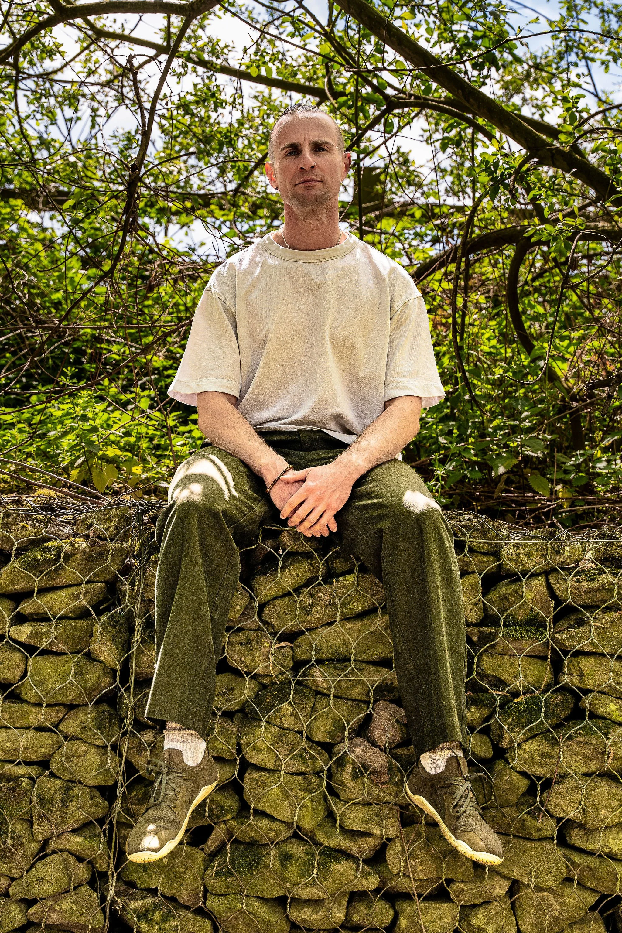 A man sits on a stone wall behind a wire fence, with green foliage and trees in the background. He is wearing a beige oversized t-shirt, baggy green pants, and sneakers.