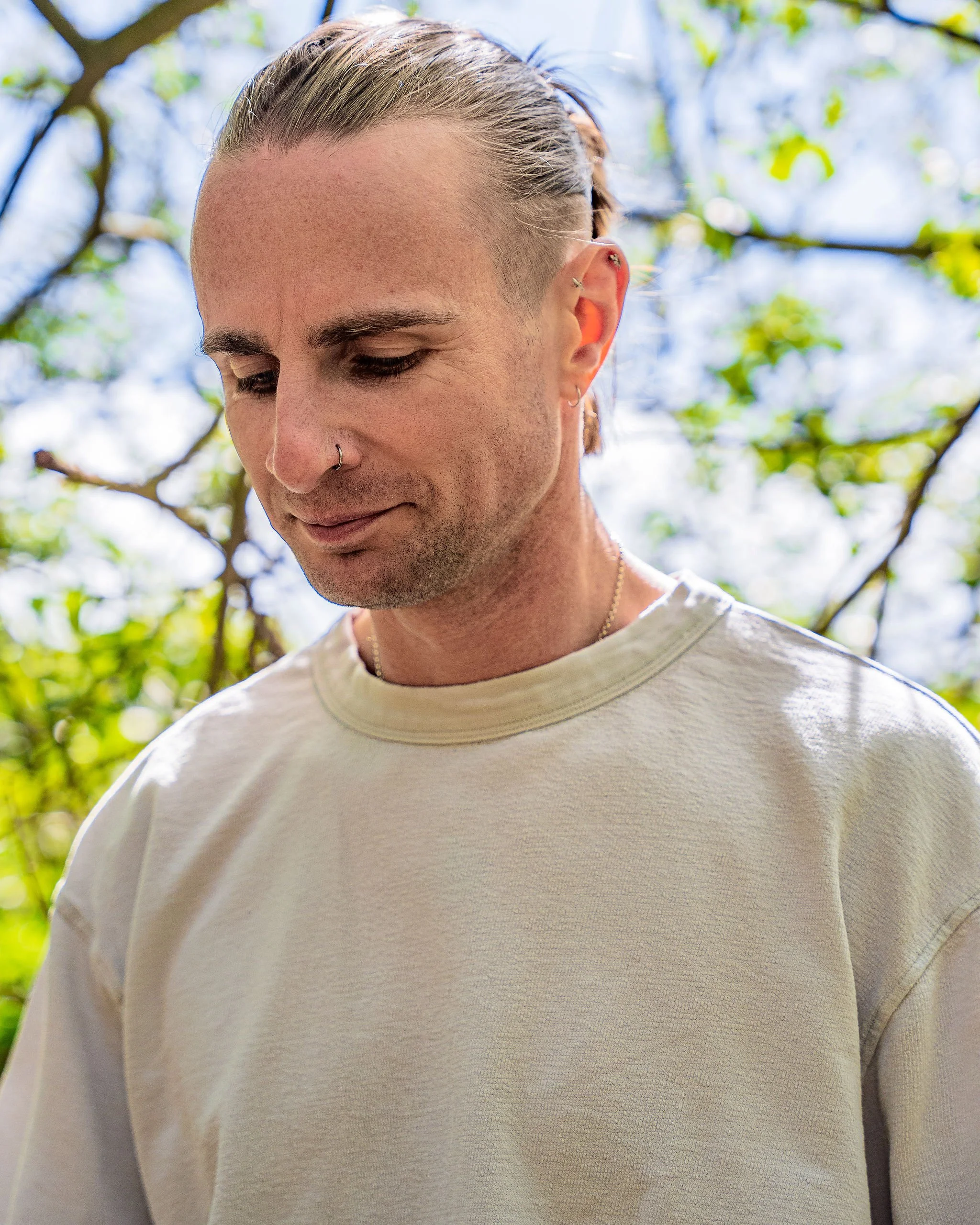 A young man with long blond hair tied back, multiple earrings, and wearing a beige sweatshirt, standing outdoors with tree leaves and sky in the background.