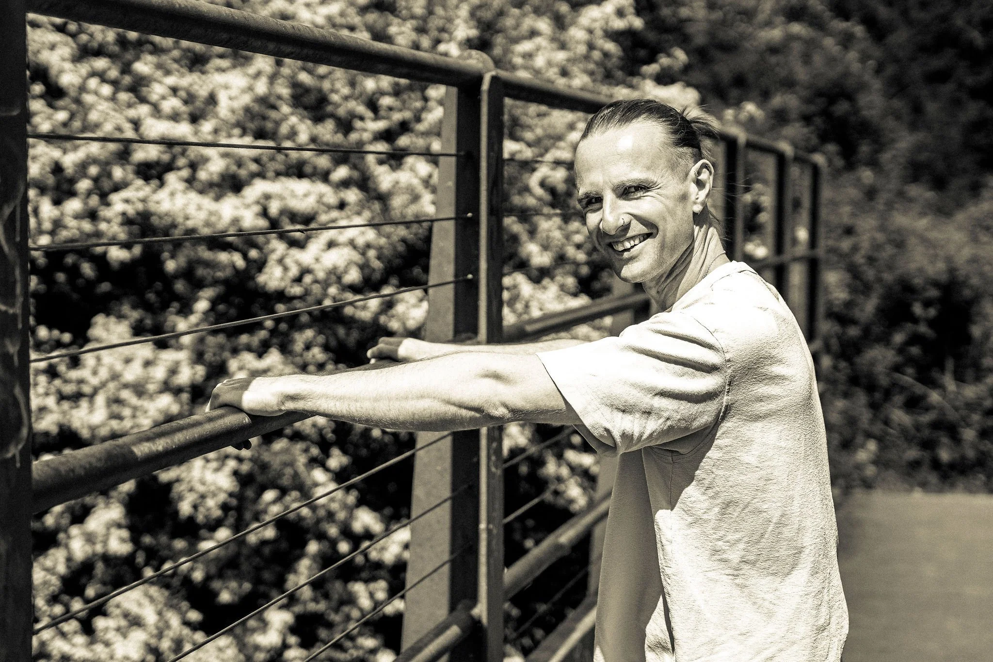 A smiling person with a shaved head and nose piercings leaning on a metal fence outdoors, with trees in the background, in black and white.