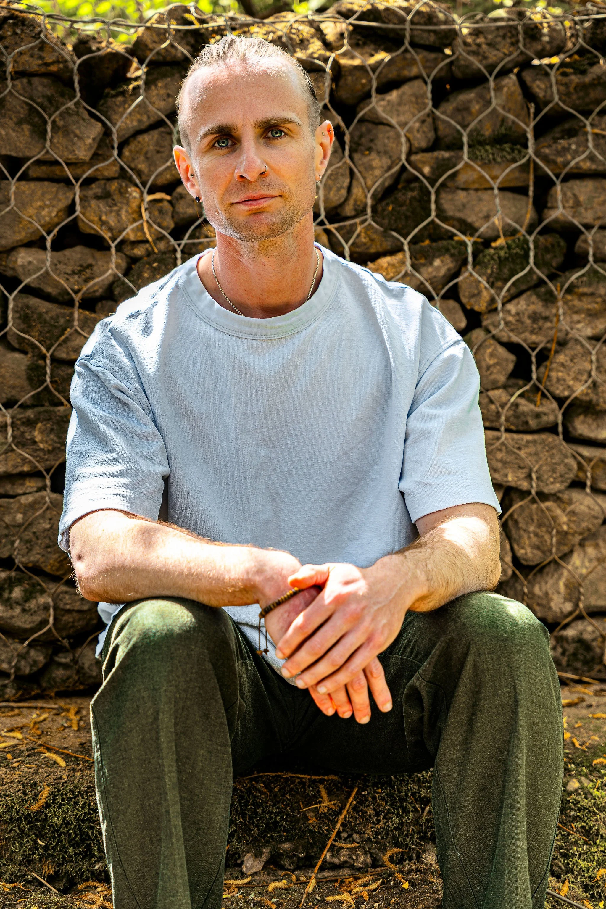 A man with blond hair and blue eyes sitting outdoors in front of a stone wall with wire fencing. He is wearing a white t-shirt and green pants, with a silver chain necklace and earrings, holding his hands together.