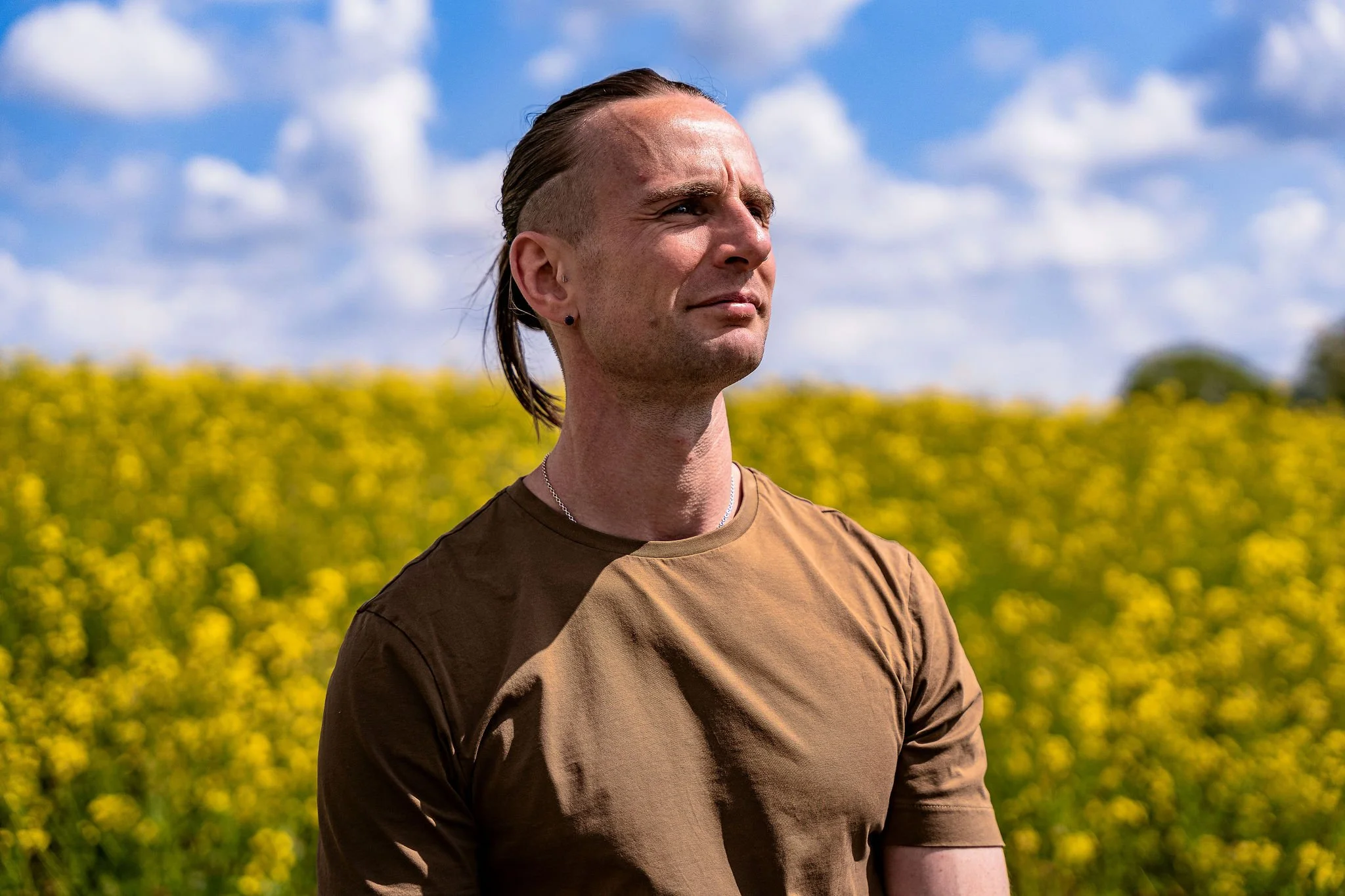 A person with styled hair, a small earring, and wearing a brown t-shirt stands outdoors in a field of yellow flowers under a partly cloudy sky.
