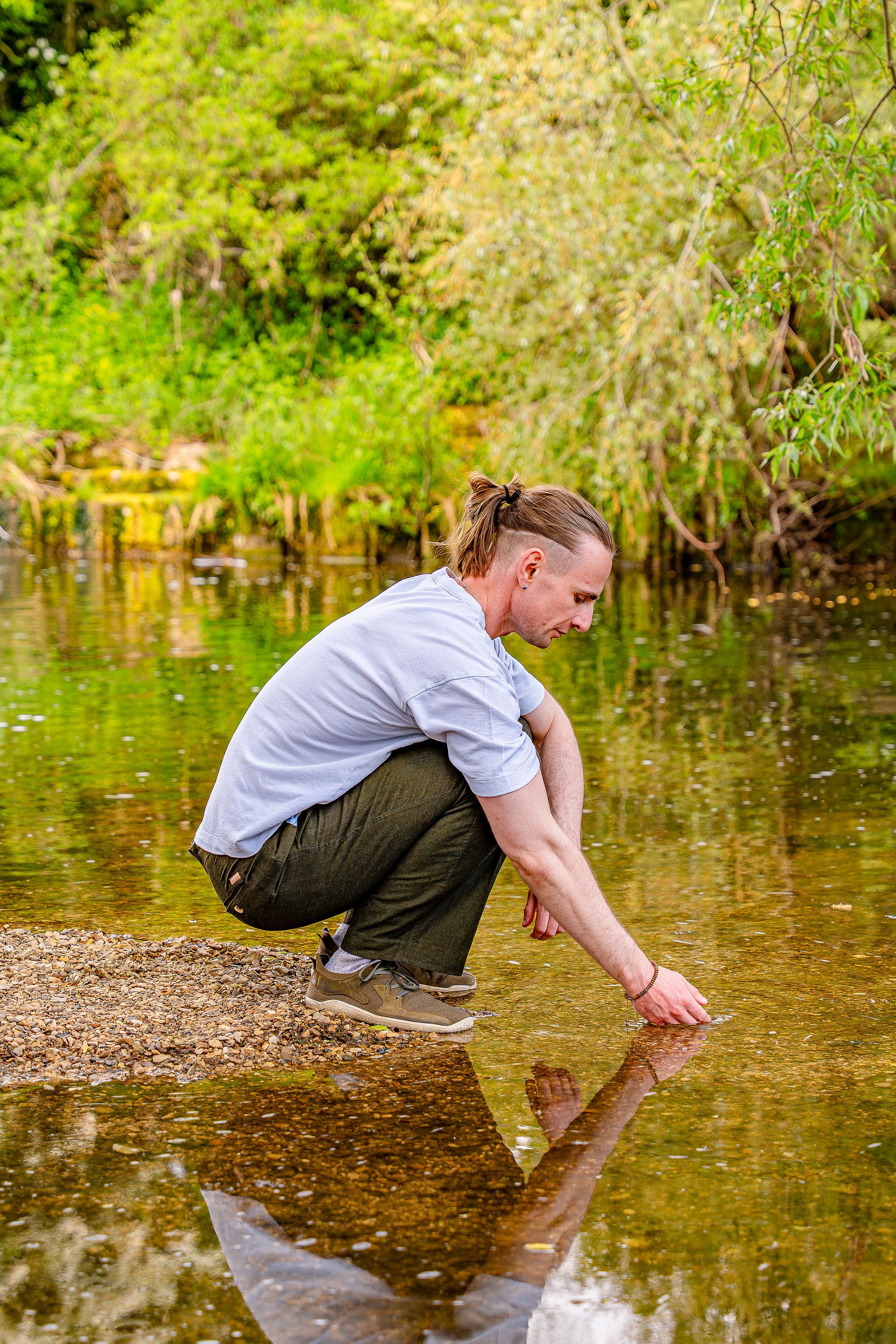 A young man with a man bun and light gray t-shirt squatting by a rocky riverbank, reaching down to touch the water, with lush green trees in the background.
