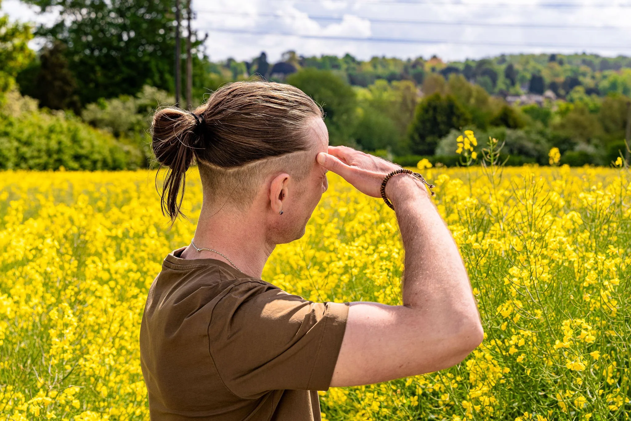 A person with short hair tied in a bun, wearing a brown shirt and bracelets, standing in a field of yellow flowers with a lush green landscape and trees in the background, saluting or shielding their eyes from the sun.