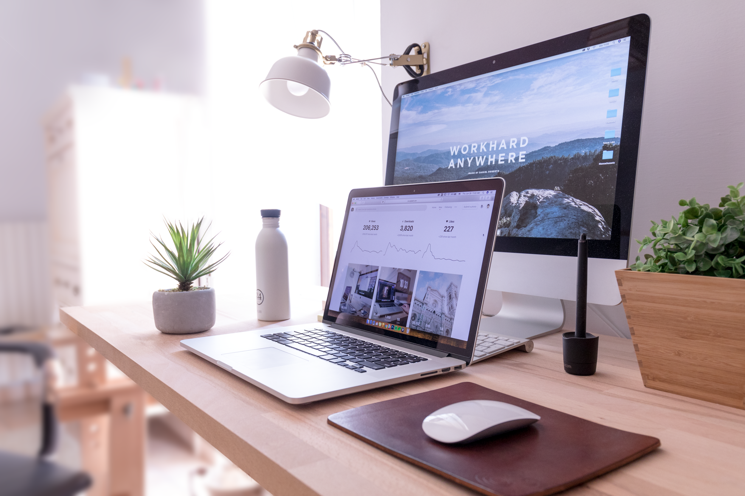 A modern workspace with a MacBook laptop, an external monitor displaying a mountain scene with the text "Workhard Anywhere," a potted green succulent, a white water bottle, and a black microphone on a wooden desk. There is a white desk lamp mounted on the wall.