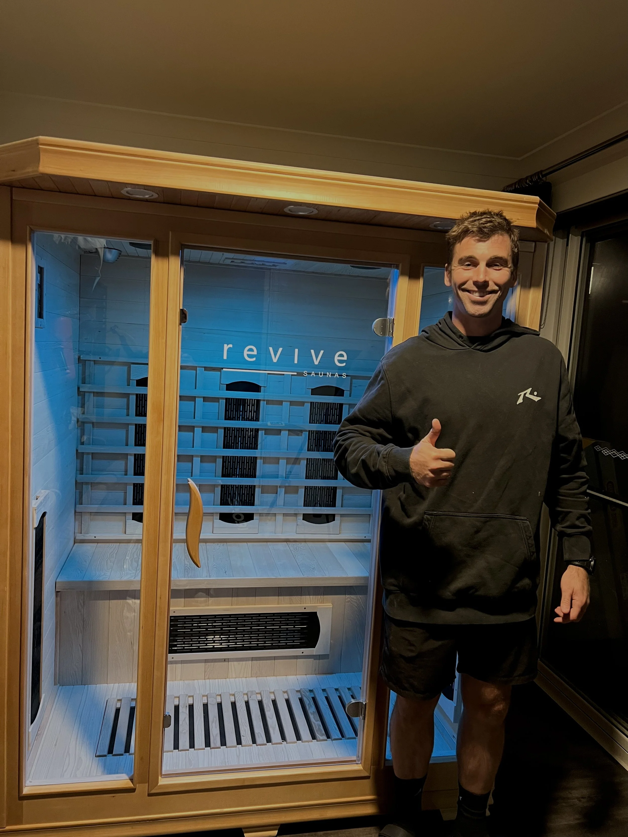 Man giving a thumbs up standing next to a wooden infrared sauna labeled "Revive Saunas" with glass doors.