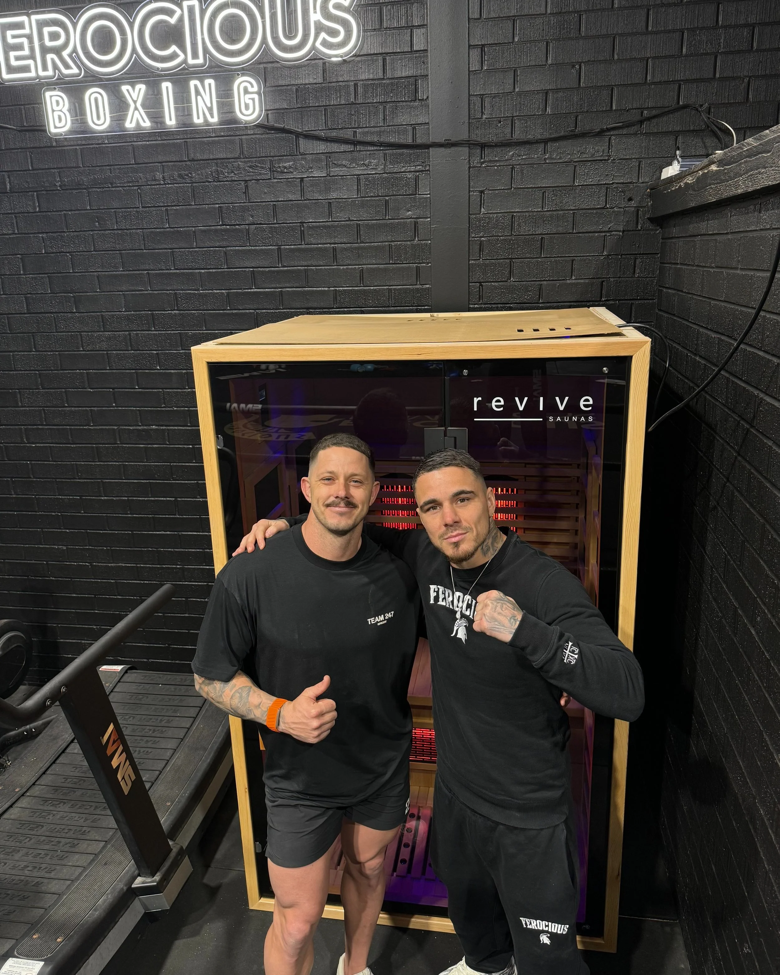 Two men posing inside a gym next to a wooden sauna. One man is wearing a 'Team Pat' t-shirt, shorts, and sneakers, giving a thumbs up. The other man is wearing 'Ferocious' branded clothing, showing a fist gesture. A treadmill labeled 'SMAI' is partially visible. The wall has 'Ferocious Boxing' signage.