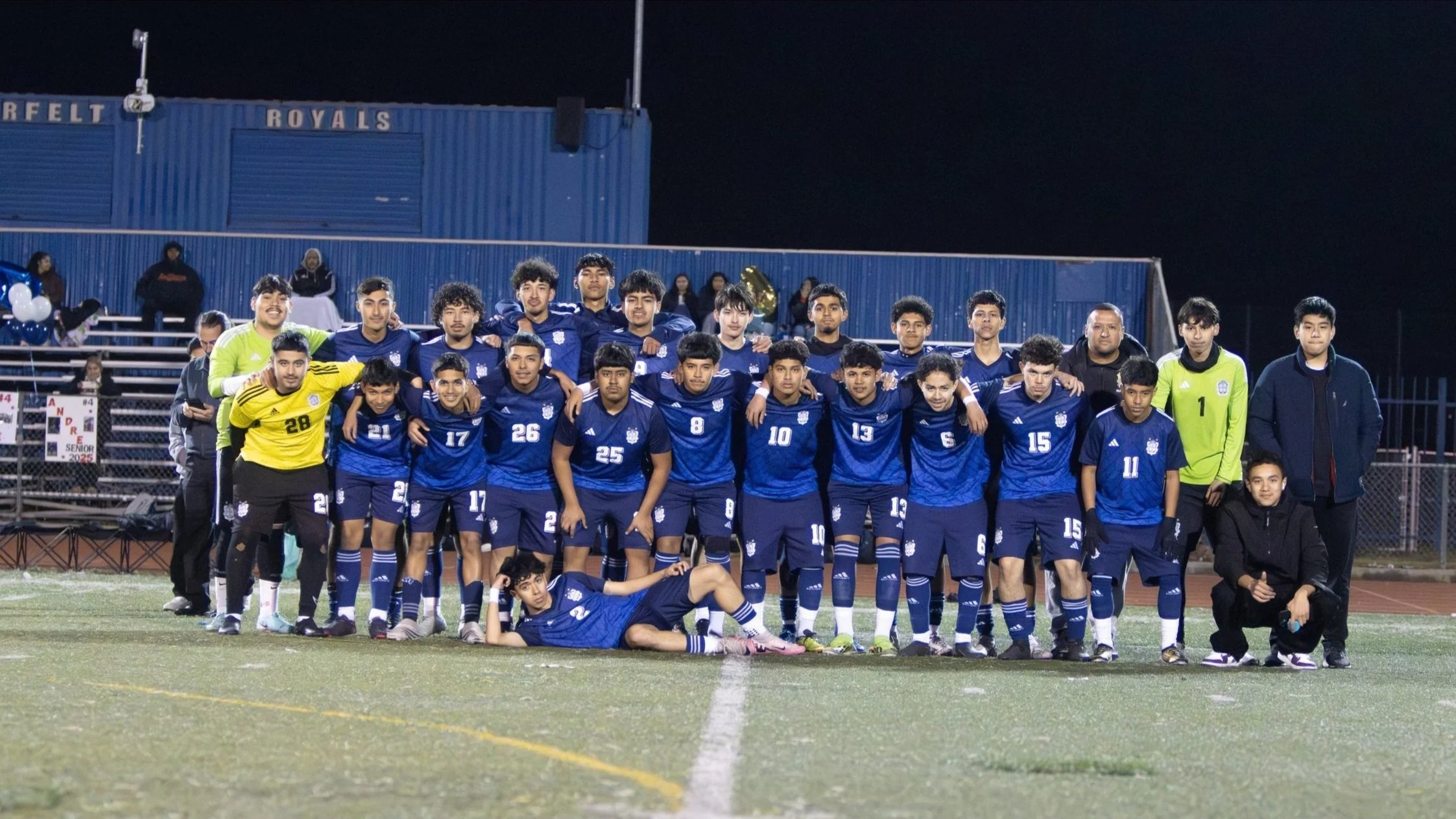 High school soccer team in blue uniforms posing on a field at night with coaches and fans in background.