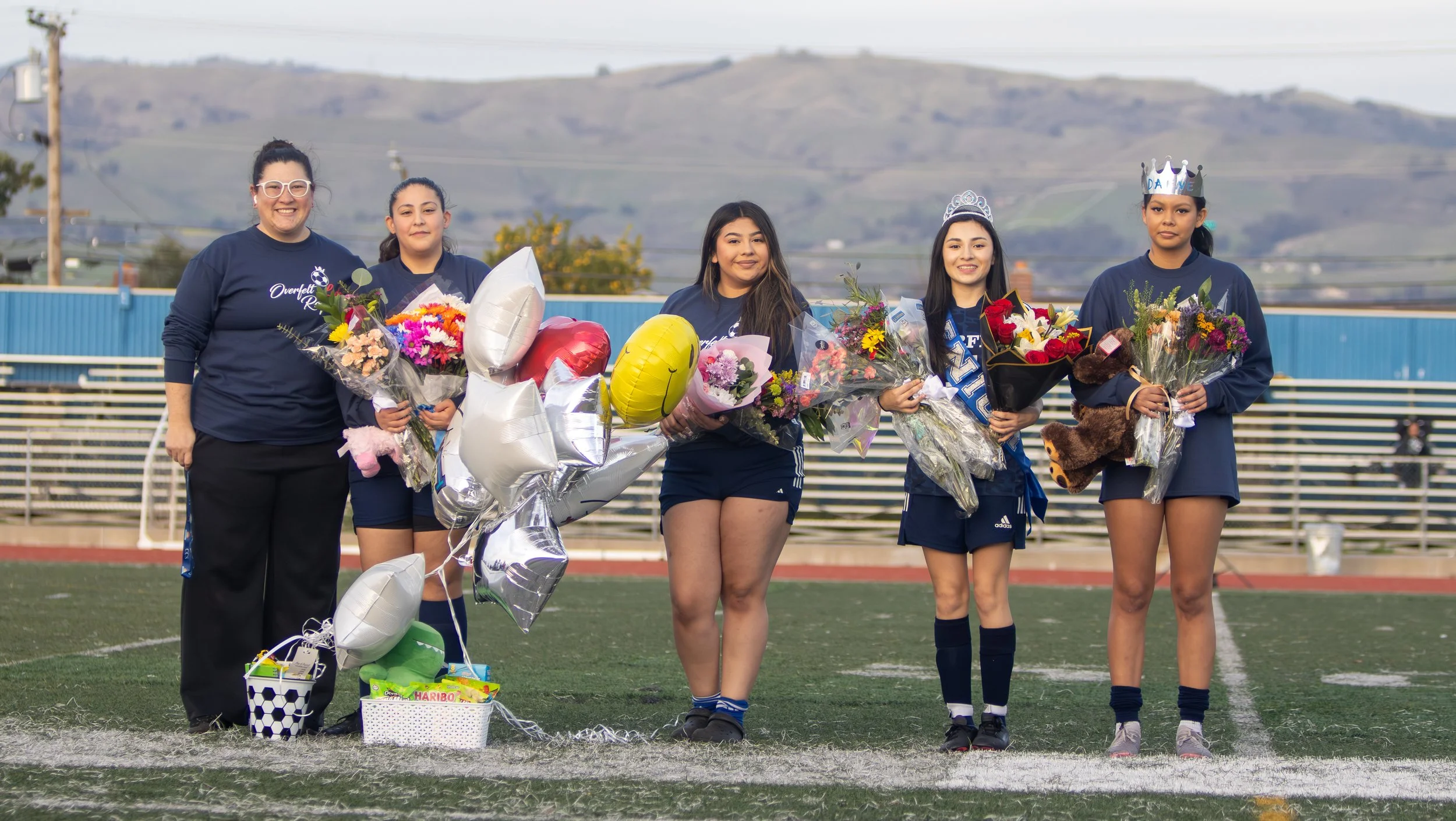 Group of women holding bouquets and balloons on a sports field, wearing blue athletic uniforms, with bleachers and hills in the background.