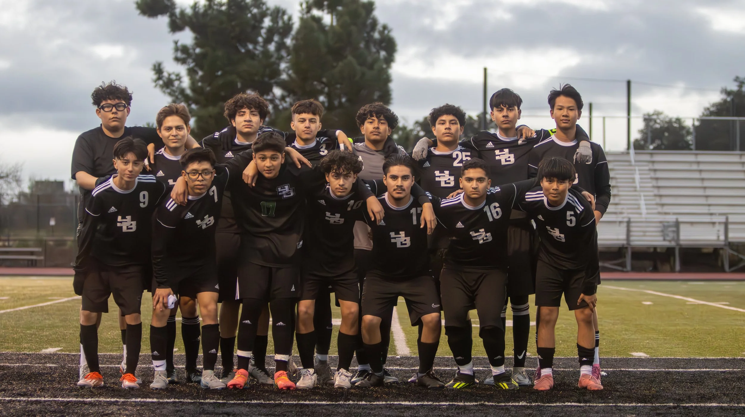 A soccer team of young males in black jerseys posing for a group photo on an outdoor field with bleachers in the background.