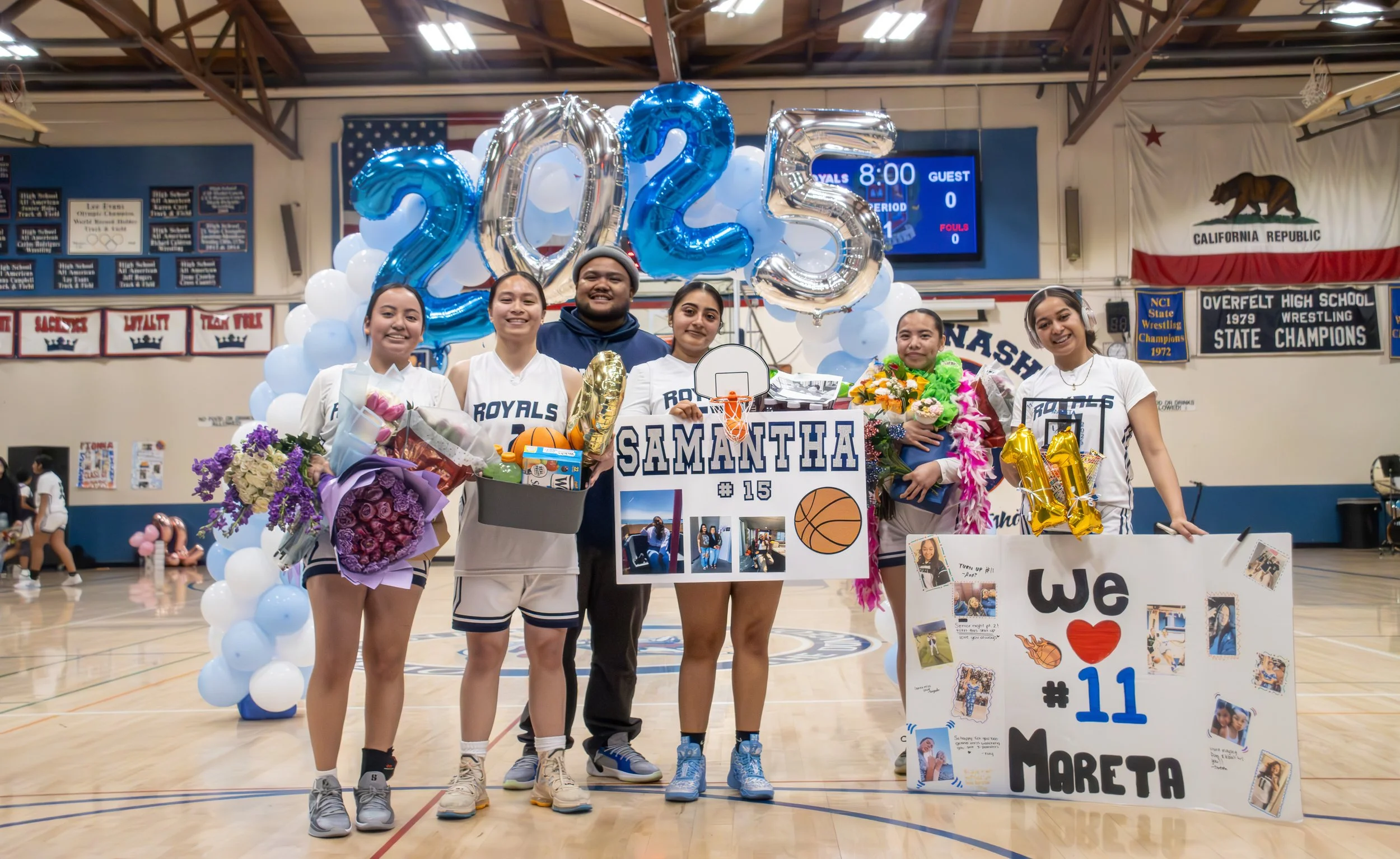 Group of young basketball players and supporters in a gym, celebrating with balloons and flowers, featuring "2025" balloons and personalized posters for team members. American flag and school banners are visible in the background.