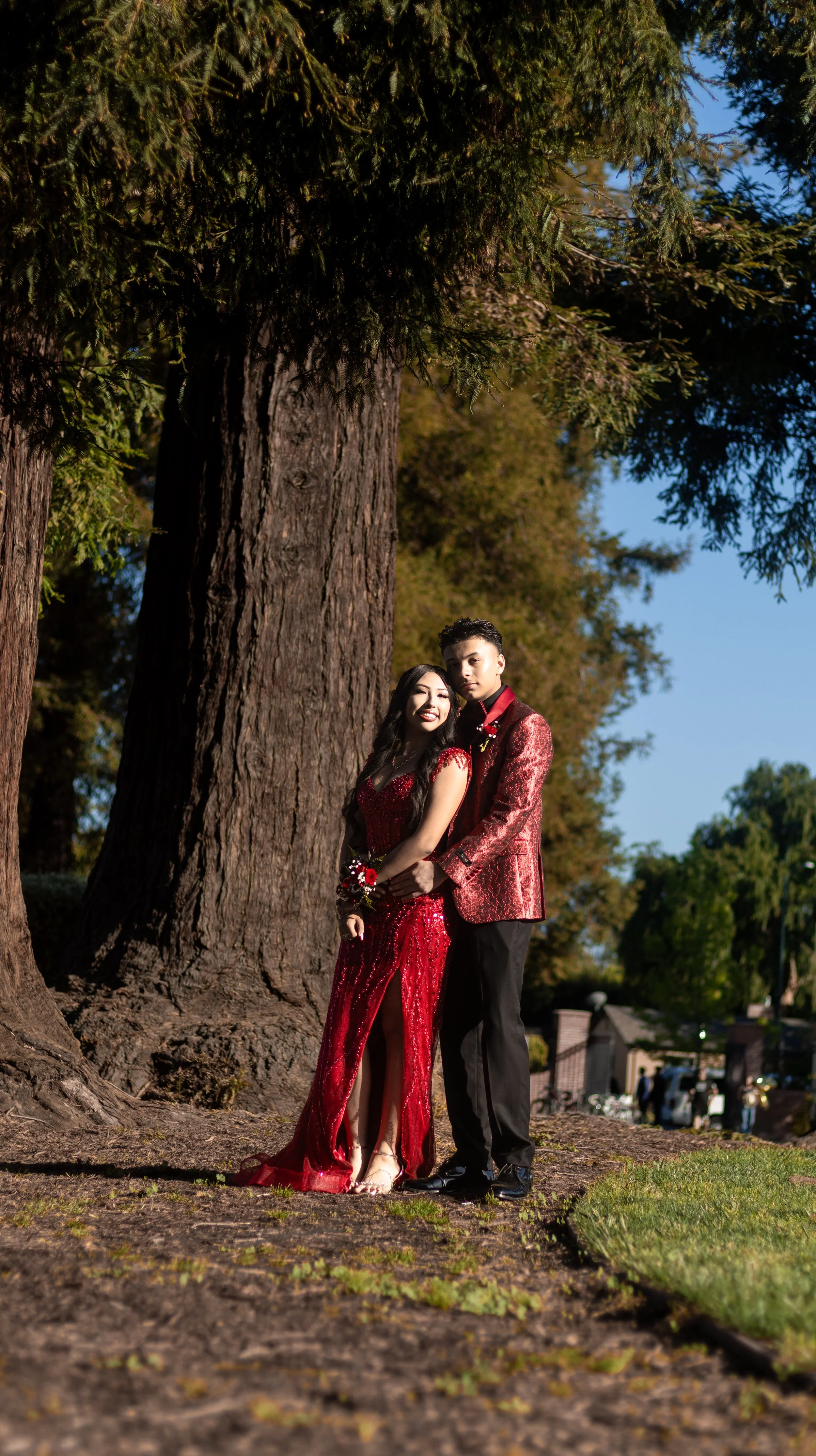 A couple dressed in matching red formal attire stands affectionately in front of a large tree in a park. The woman is wearing a red sequined gown, and the man is in a red patterned jacket. The setting is a sunlit outdoor area with greenery and a clear blue sky.