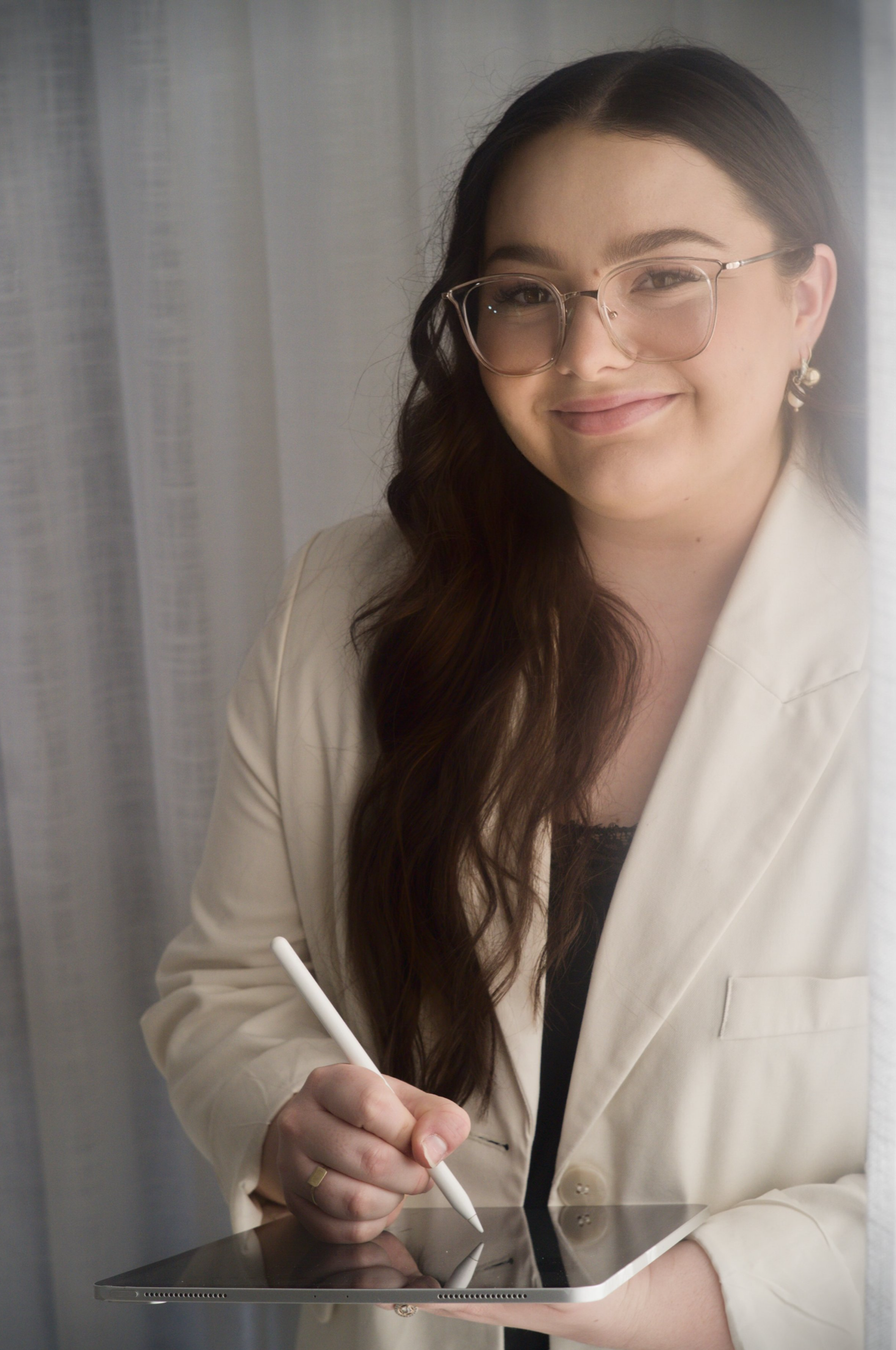 A woman with long brown hair, glasses, and earrings, smiling while using a stylus on a tablet.