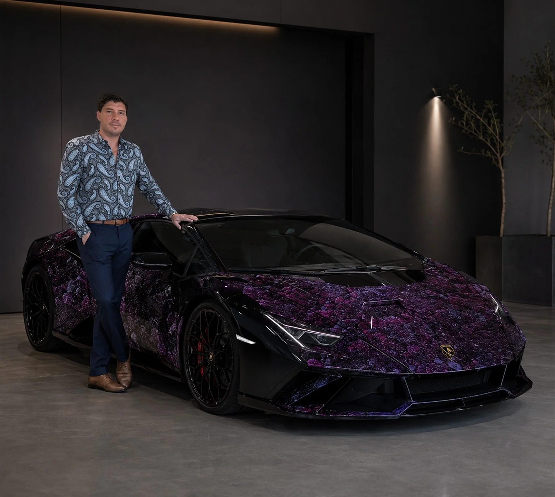 A man dressed in a paisley patterned shirt and navy pants standing next to a purple camouflage Lamborghini sports car in a modern indoor showroom with dark walls and minimal lighting.