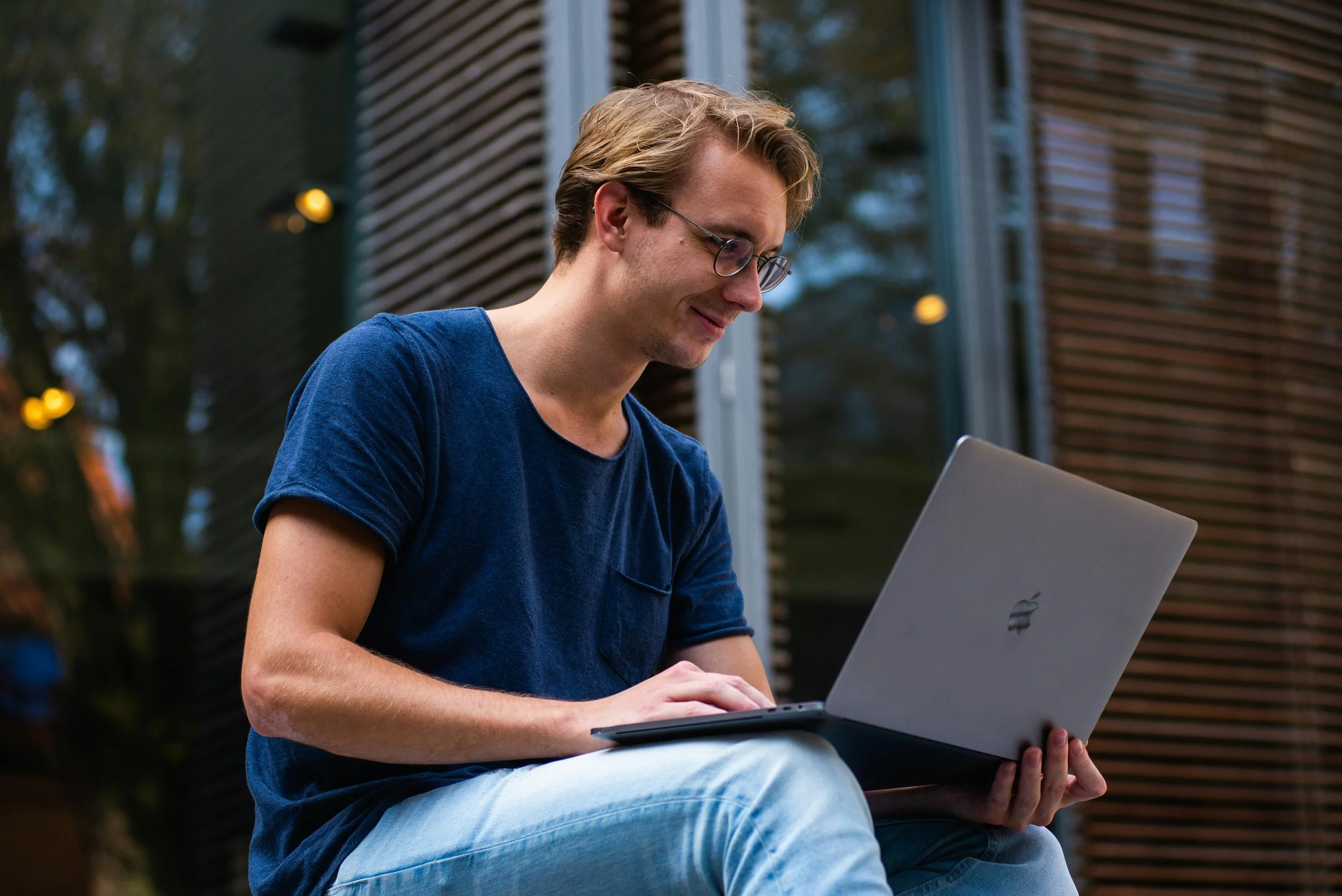 A young man with glasses and blond hair wearing a blue T-shirt and light blue jeans, smiling while working on a silver MacBook outdoors in front of a modern building.