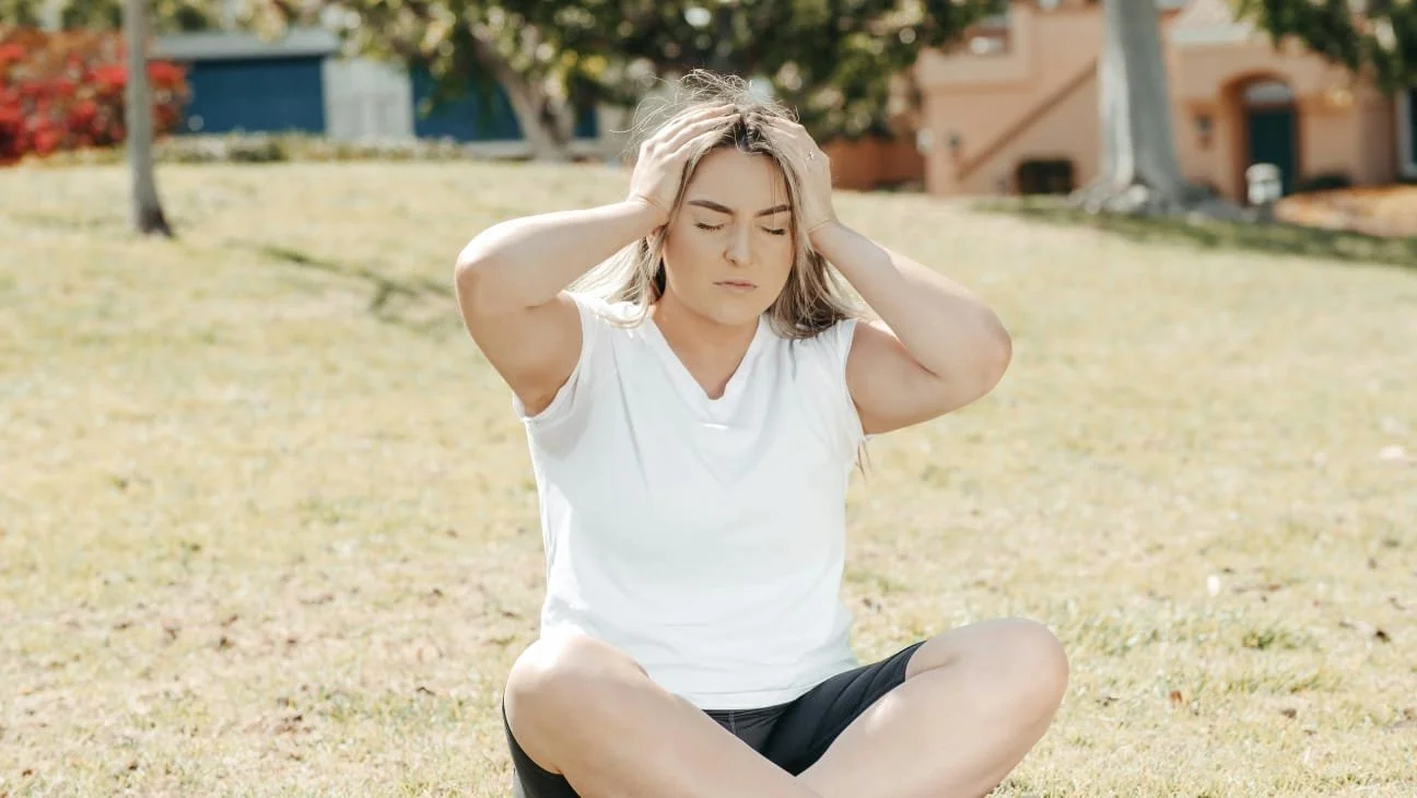 Woman sitting outdoors on grass, holding her head, appearing distressed or worried.