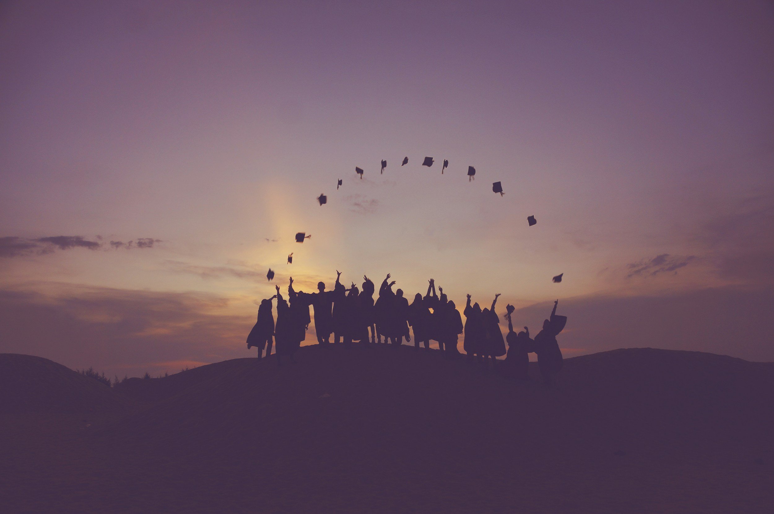 A group of graduates in caps and gowns celebrating on a hilltop at sunset, throwing their caps into the air.