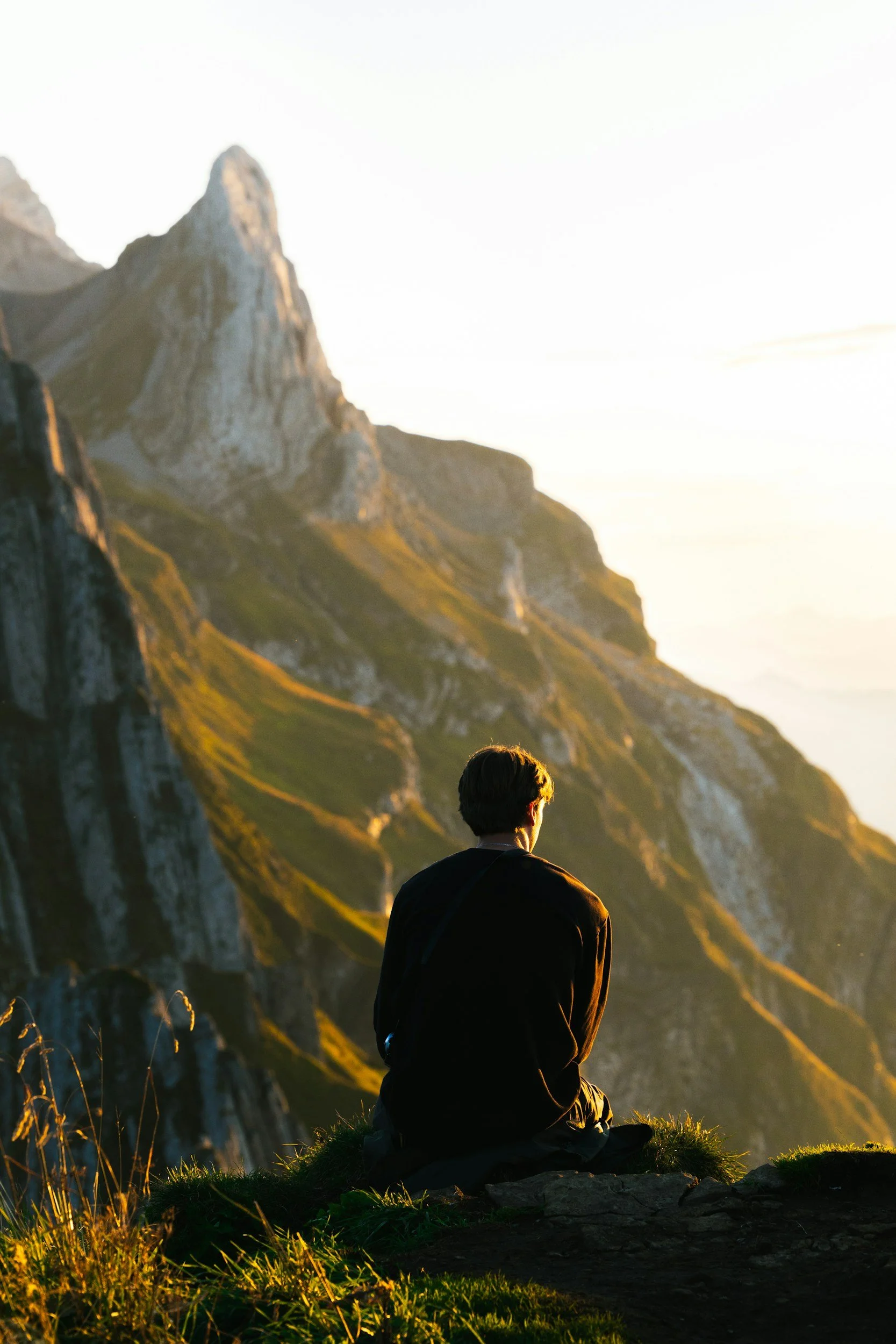 A person sitting on a grassy hill, facing away, observing a mountain landscape during sunset.