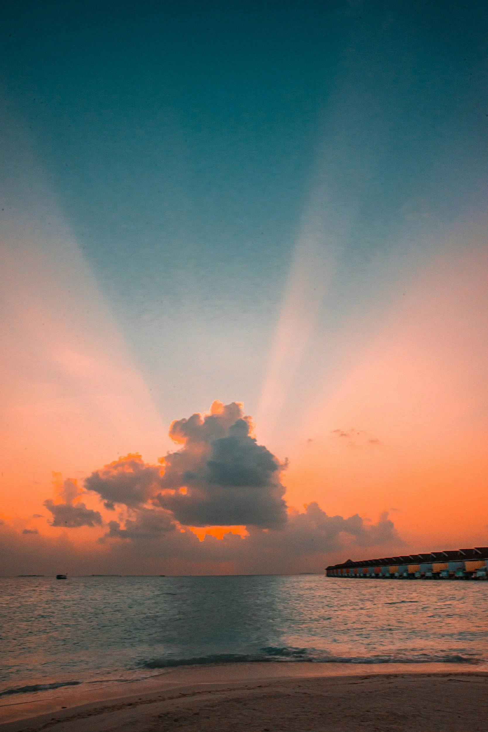 Sunset over the ocean with clouds in the sky, a small boat in the water, and overwater bungalows along the shoreline.
