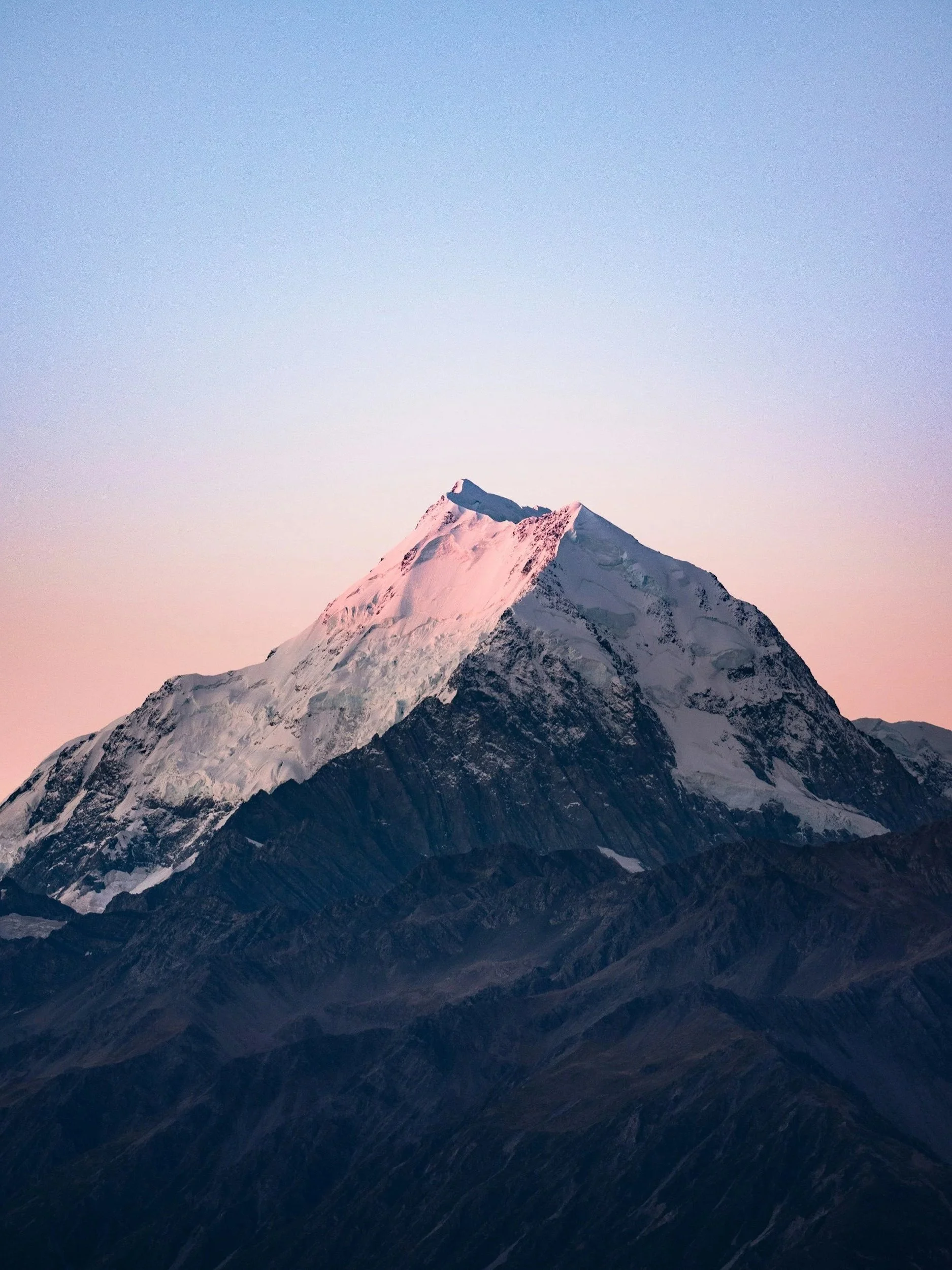Snow-covered mountain peak at sunset with pinkish light on the summit and a clear sky in the background.