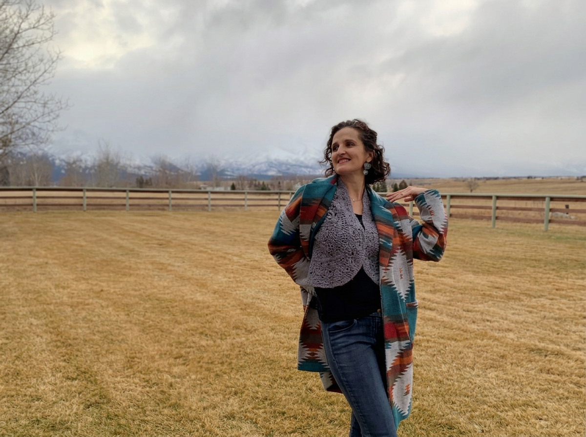 A woman with curly hair smiling while standing on a grassy field with a fence behind her and mountains in the distance, wearing a colorful patterned coat and earrings.
