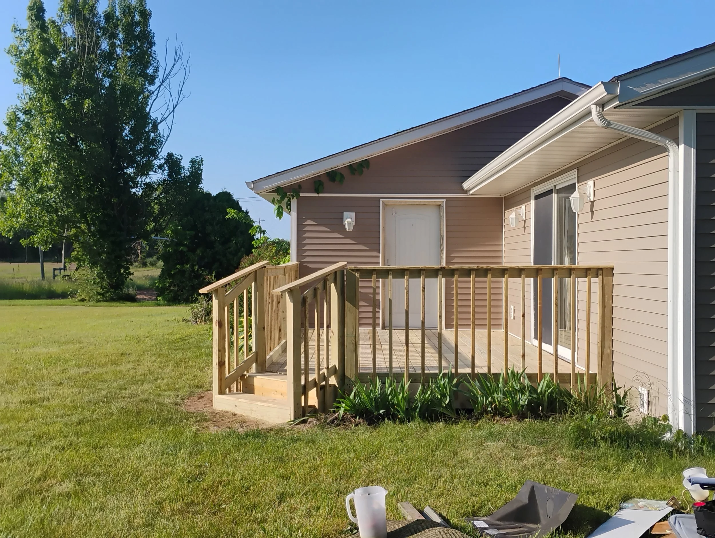 Back porch with railing and security door on a beige house, surrounded by green lawn and trees, under a blue sky.