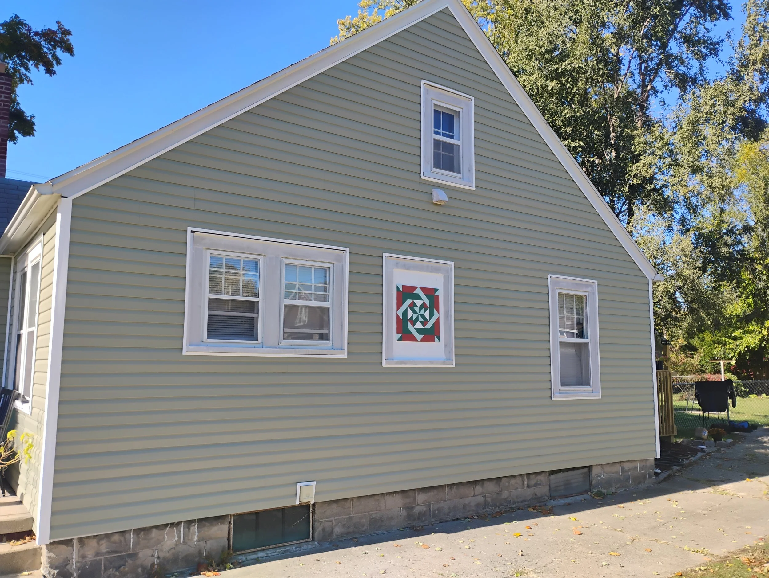 Side view of a green house with white trim, three windows, and a geometric quilt pattern on the wall, surrounded by trees and a yard.