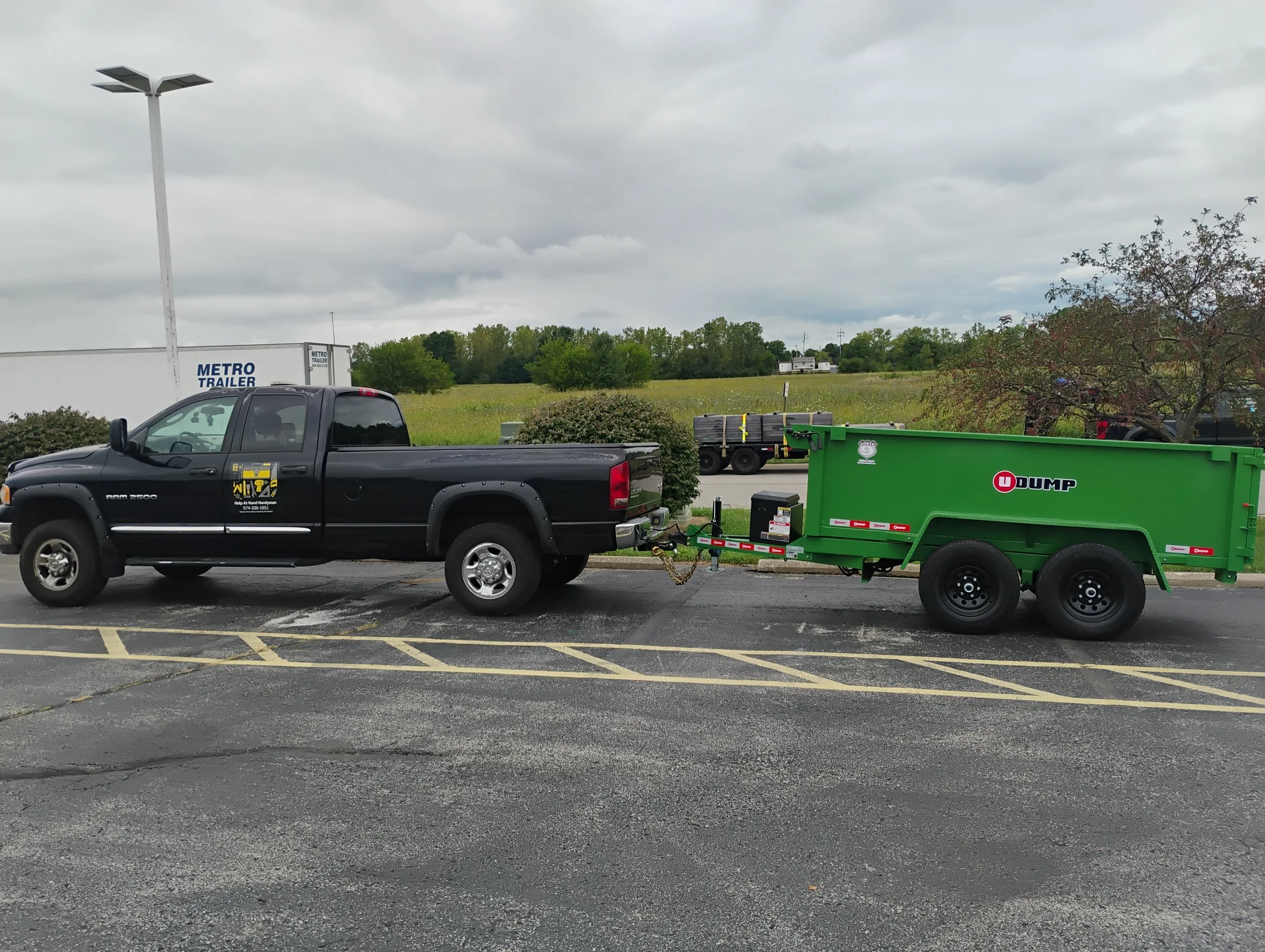A black pickup truck with a trailer attached parked in a lot on a cloudy day. The trailer is green and labeled with a red and black logo, and it is linked to the truck by a chain. In the background, there are open fields, trees, and some stacked cargo on a smaller trailer.