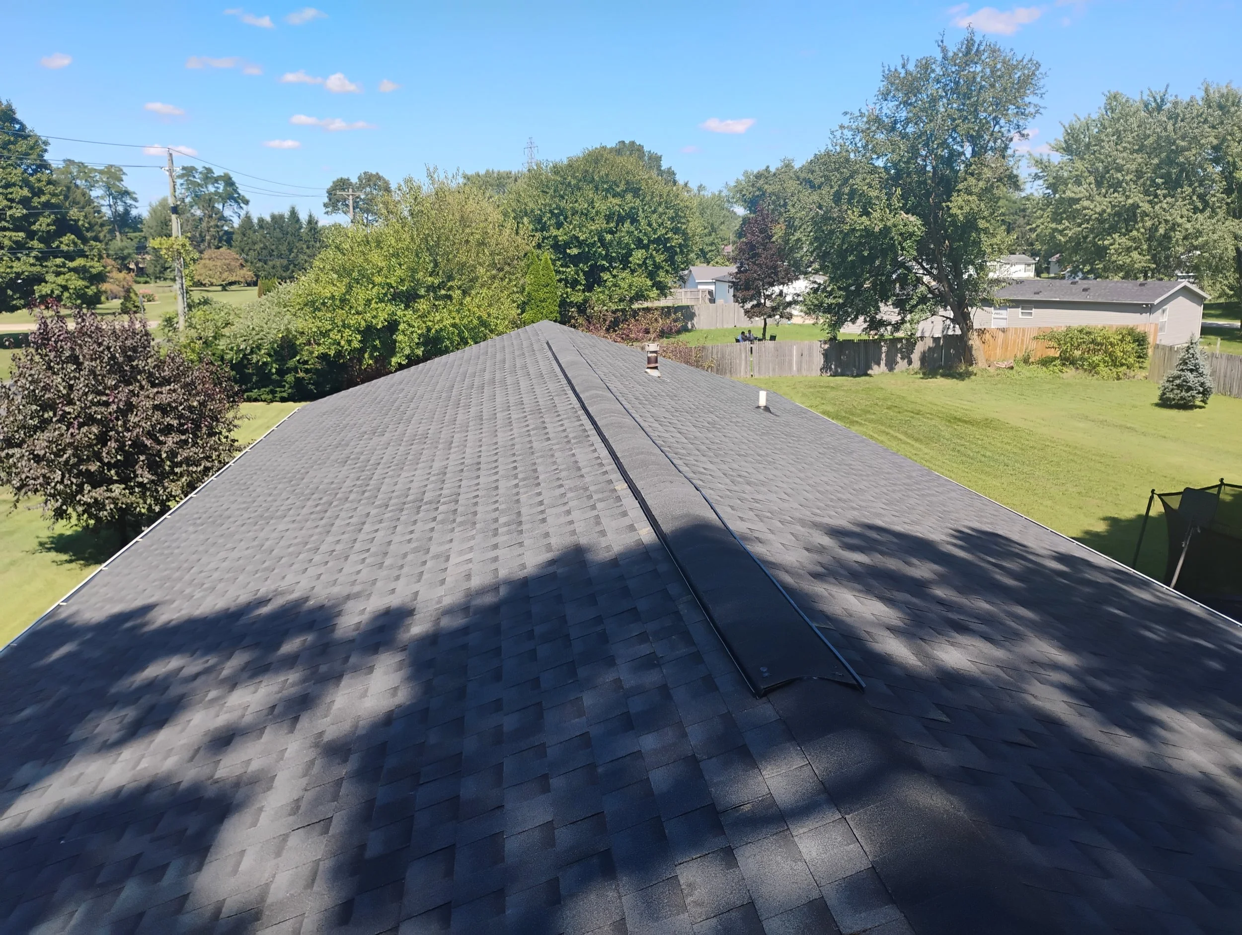 View of a residential roof with gray shingles, small vent pipes, surrounded by trees and a fenced backyard under a clear blue sky.