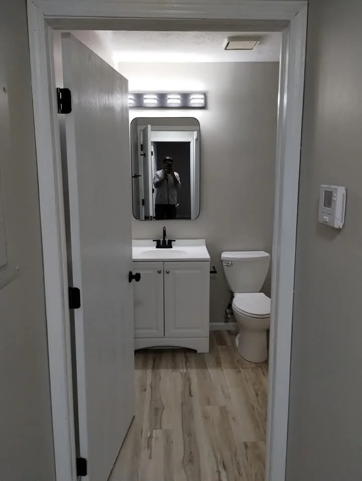 A small bathroom with white walls, a white vanity with a black faucet, a mirror above it, and a toilet to the right. There is a light fixture above the mirror and a thermostat or control panel on the wall to the right.