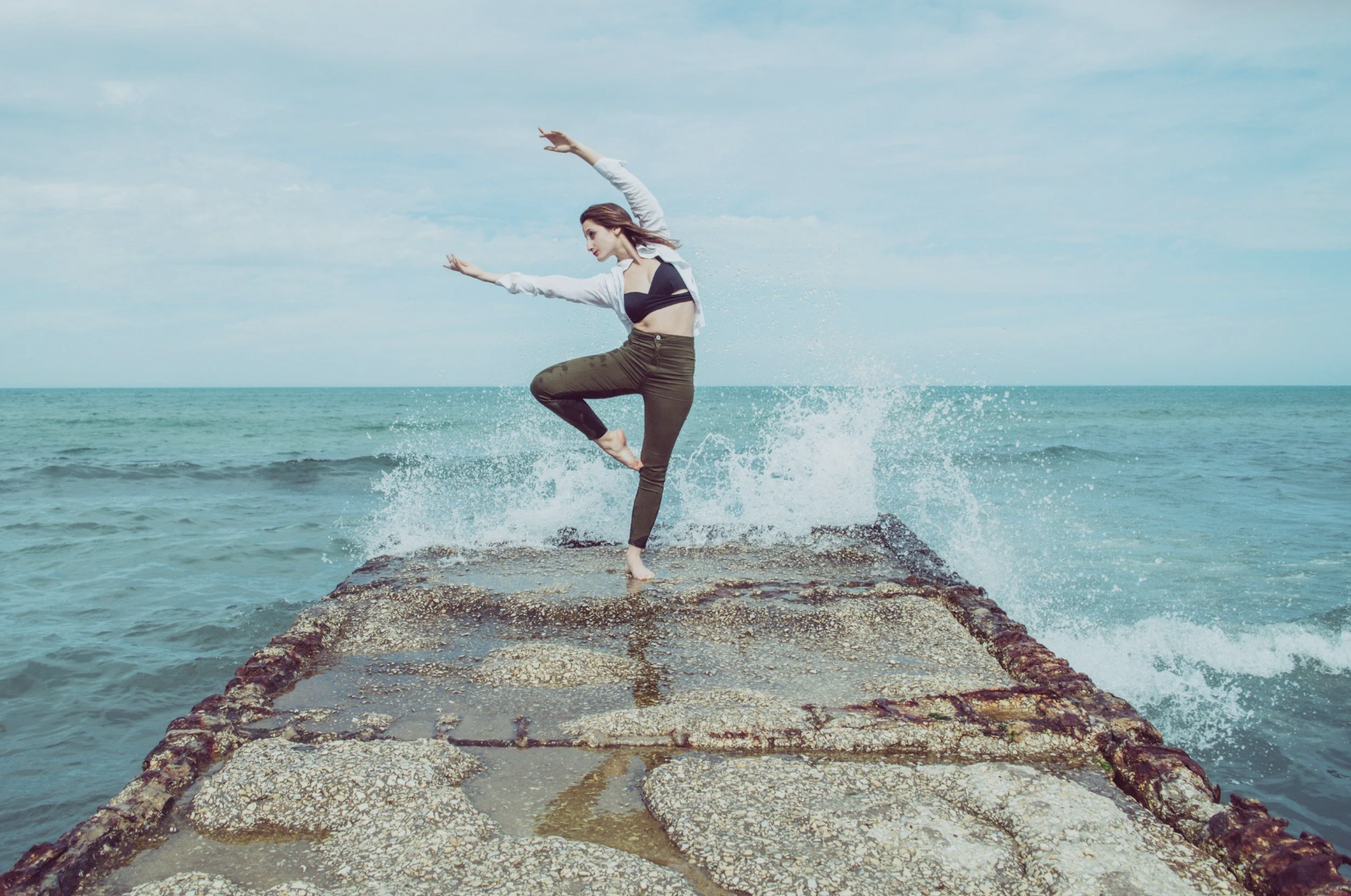 A woman dancing on a rock pier by the ocean, with waves splashing around her.