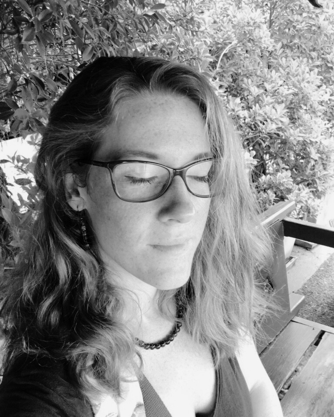 A woman with long wavy hair and glasses with closed eyes, sitting outdoors on a bench, surrounded by foliage.