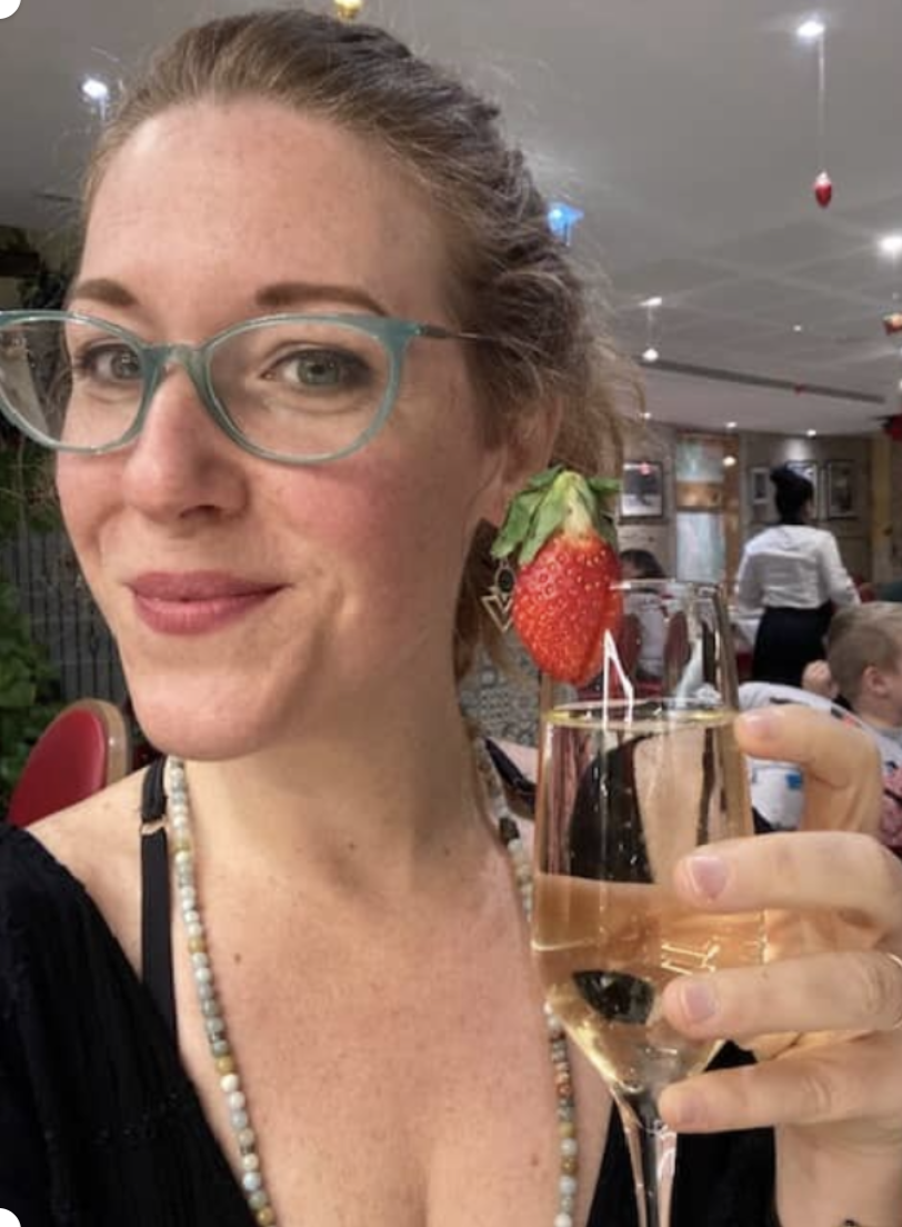woman with glasses holding a glass of champagne with a strawberry garnish, in a restaurant or party setting.