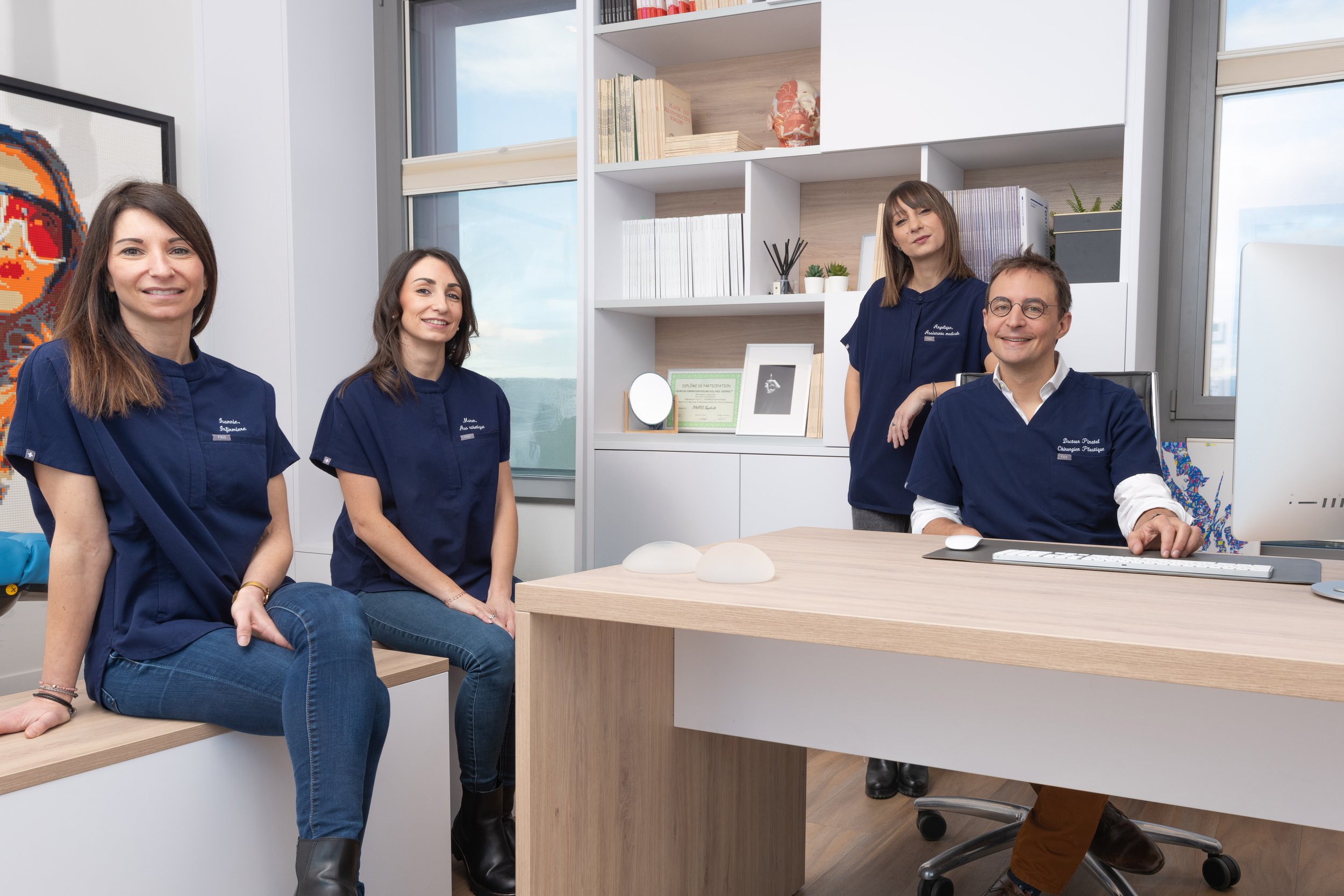 Groupe de cinq professionnels dans un bureau moderne, portant des blouses bleues, souriants, autour d'une table en bois, avec des étagères et des décorations visibles en arrière-plan.