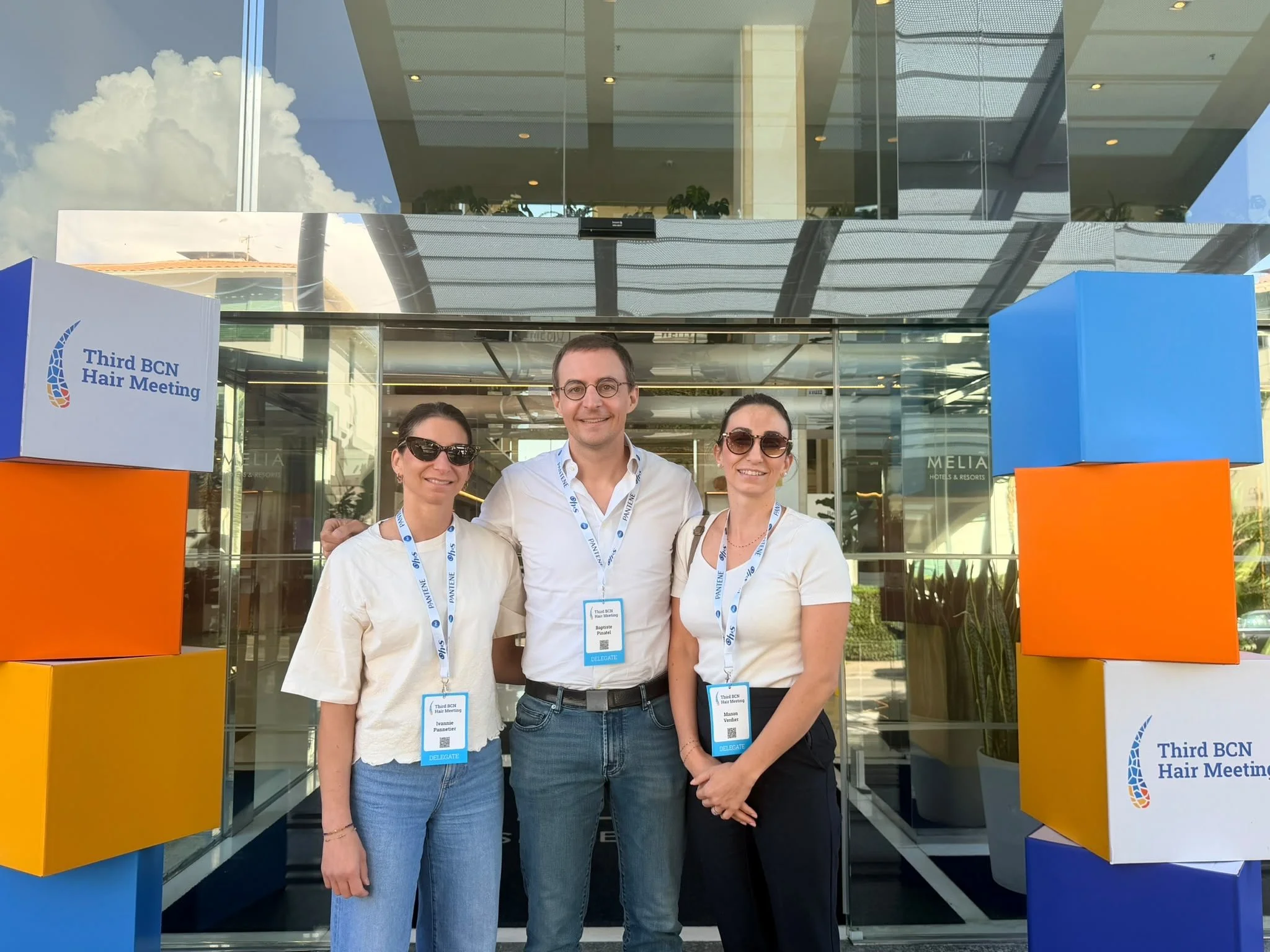 Trois personnes posent pour une photo devant l'entrée d'un lieu d'événement, avec des panneaux indiquant « Third BCN Hair Meeting » et des badges nominaux pendus autour du cou.