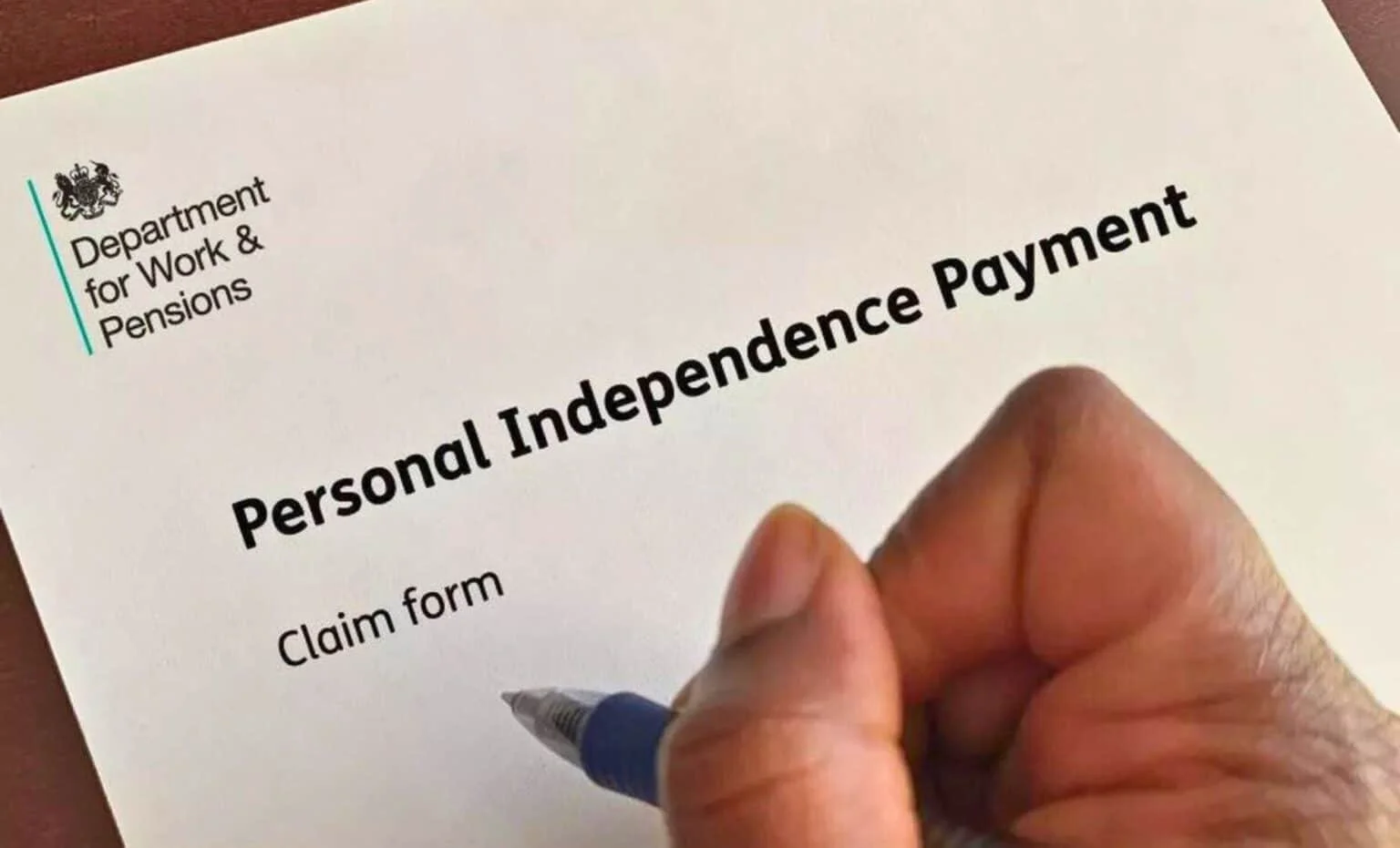 Close-up of a person holding a pen over a "Personal Independence Payment" claim form from the Department for Work & Pensions.