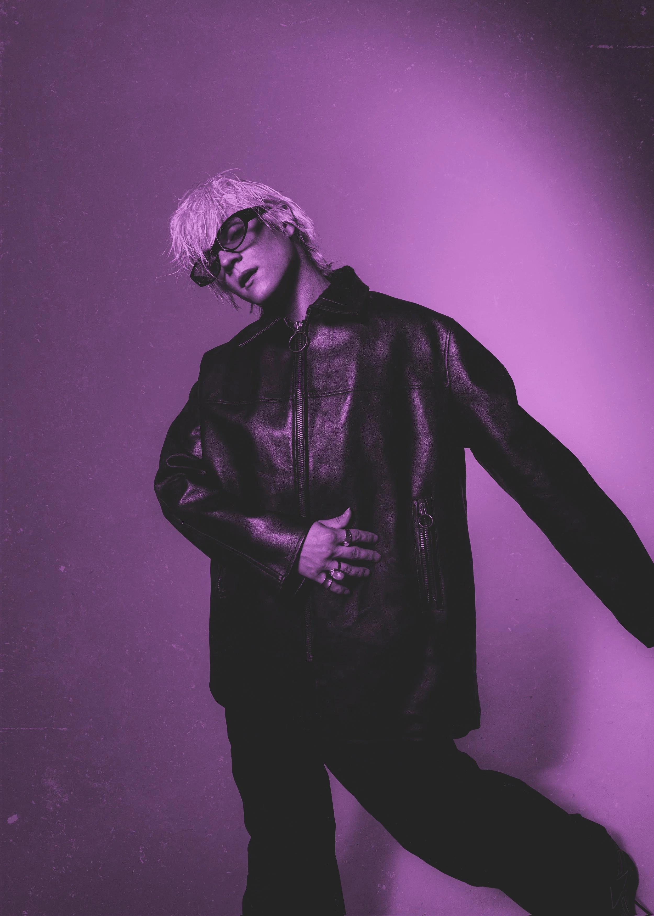 Person with blond hair, glasses, and black leather jacket standing against purple background.
