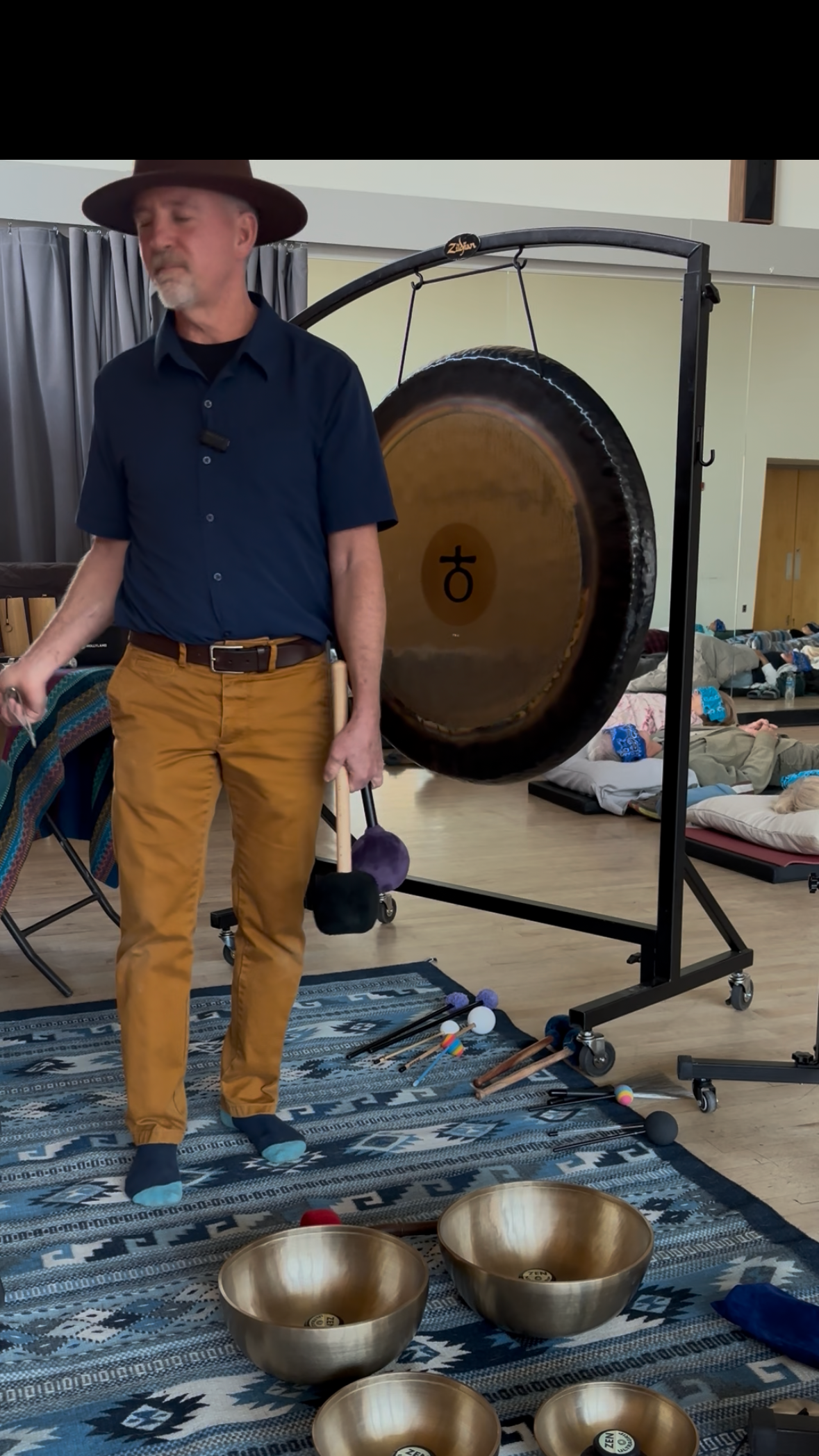 A man wearing a wide-brimmed hat, a navy blue shirt, and mustard-colored pants stands in front of a large gong with a symbol on its surface. The gong is on a black metal stand, and the man holds a mallet in his right hand. In front of him are several singing bowls and small mallets on a patterned rug. Behind him, there are people lying on mats on the floor, some with blue sock-like covers on their feet, suggesting a relaxation or meditation session.