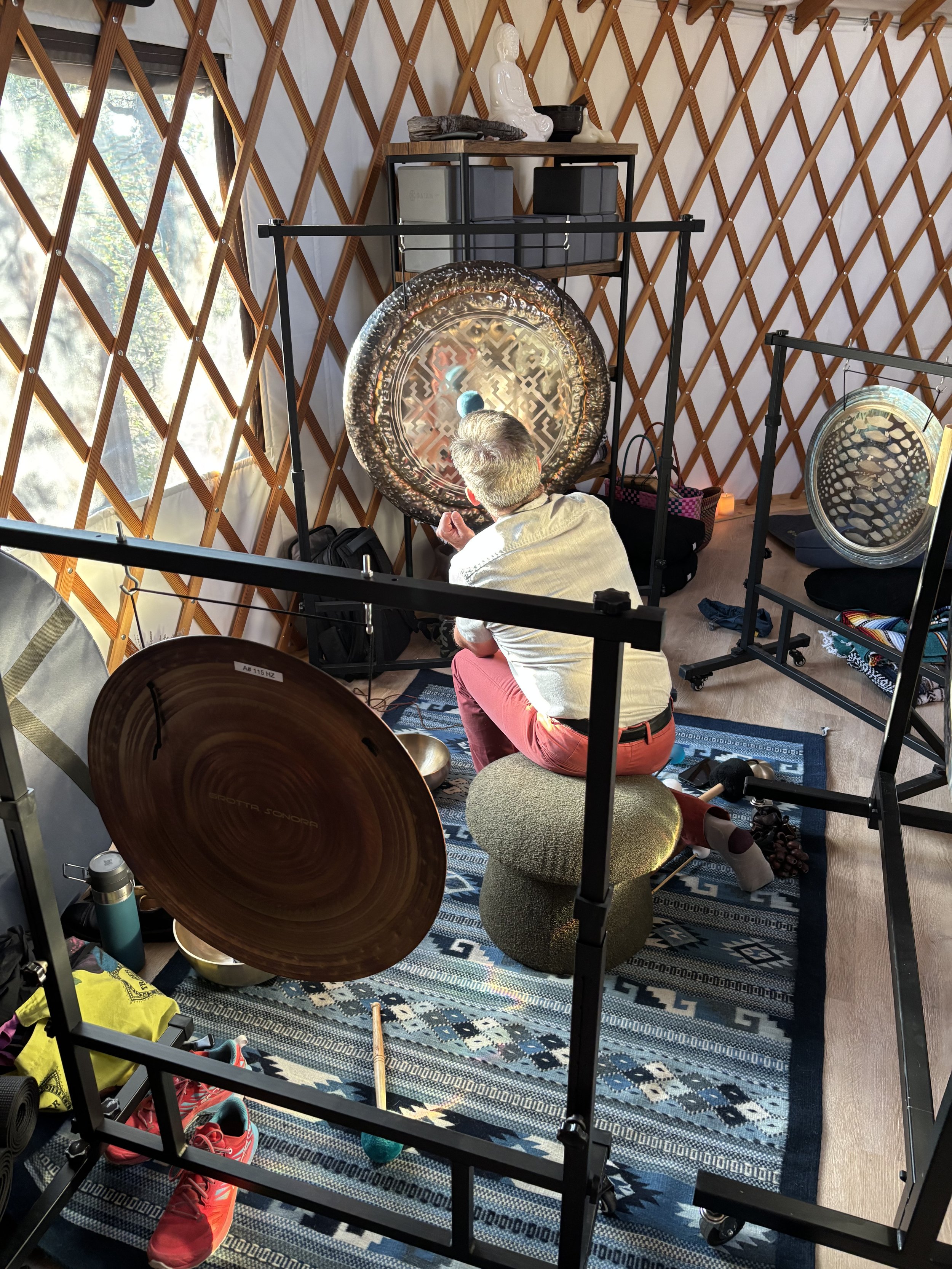 A person is sitting on a green cushion, playing a gong with a mallet inside a yurt with a wooden lattice wall. The person's back is turned, and they are wearing a beige shirt and pink pants. There are various musical instruments and personal items around, including a cymbal, shoes, water bottle, and bags.