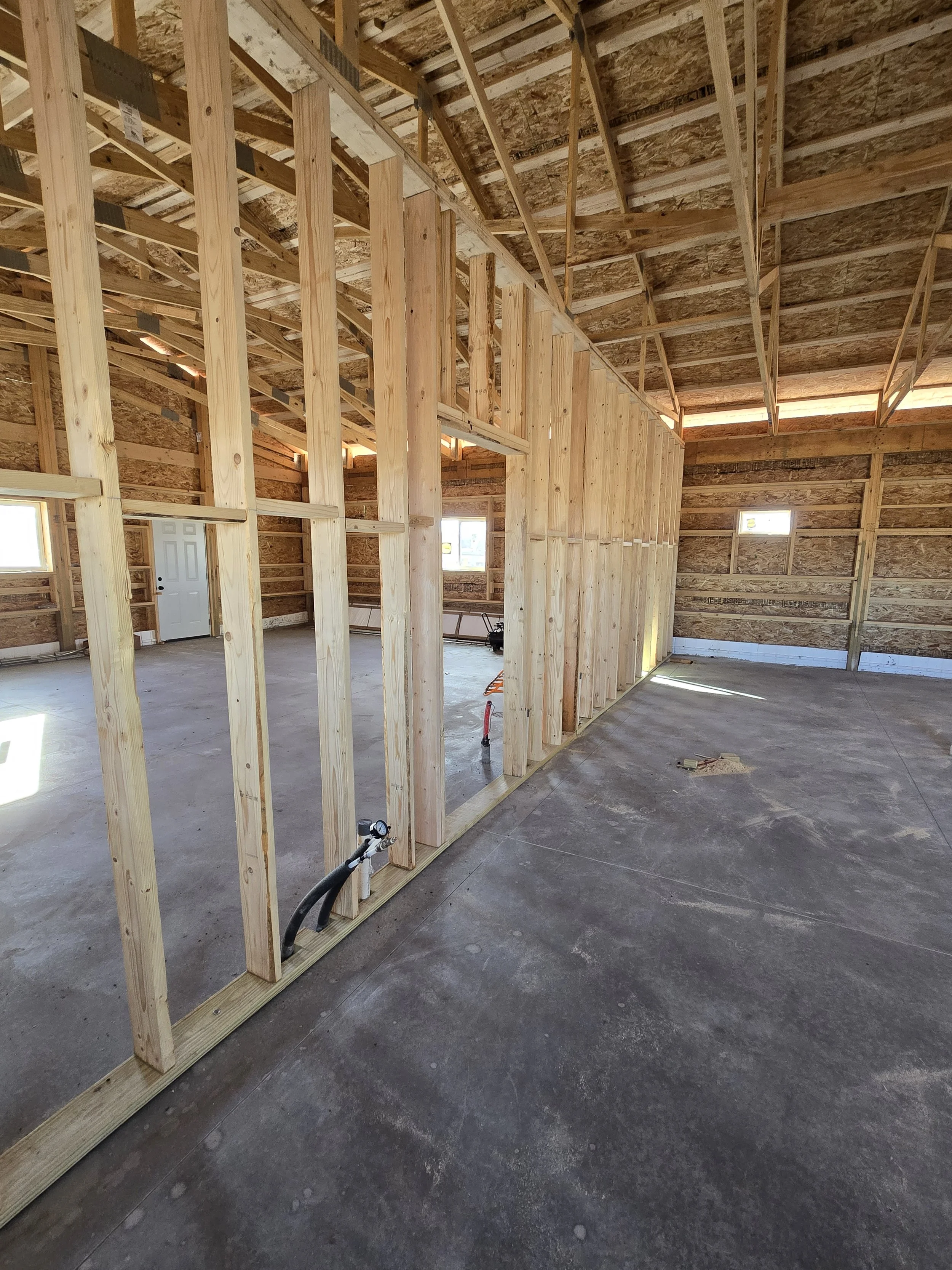Interior of a house under construction with exposed wooden framing, a concrete floor, small windows, and unfinished walls.
