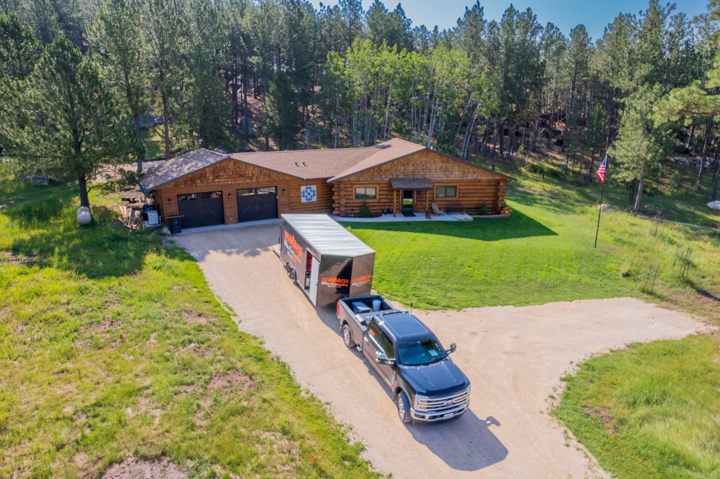 A house with a wooden exterior surrounded by trees, a grassy lawn, a driveway with a pickup truck, a moving truck, and an American flag on a pole.