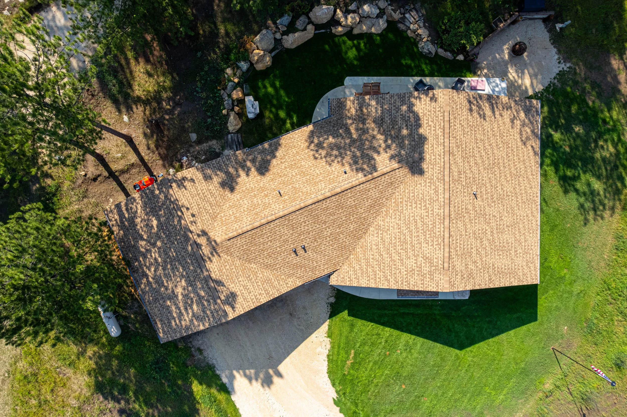 An aerial view of a house with a brown shingle roof, surrounded by trees, grass, a gravel driveway, and outdoor furniture including chairs and a fire pit.