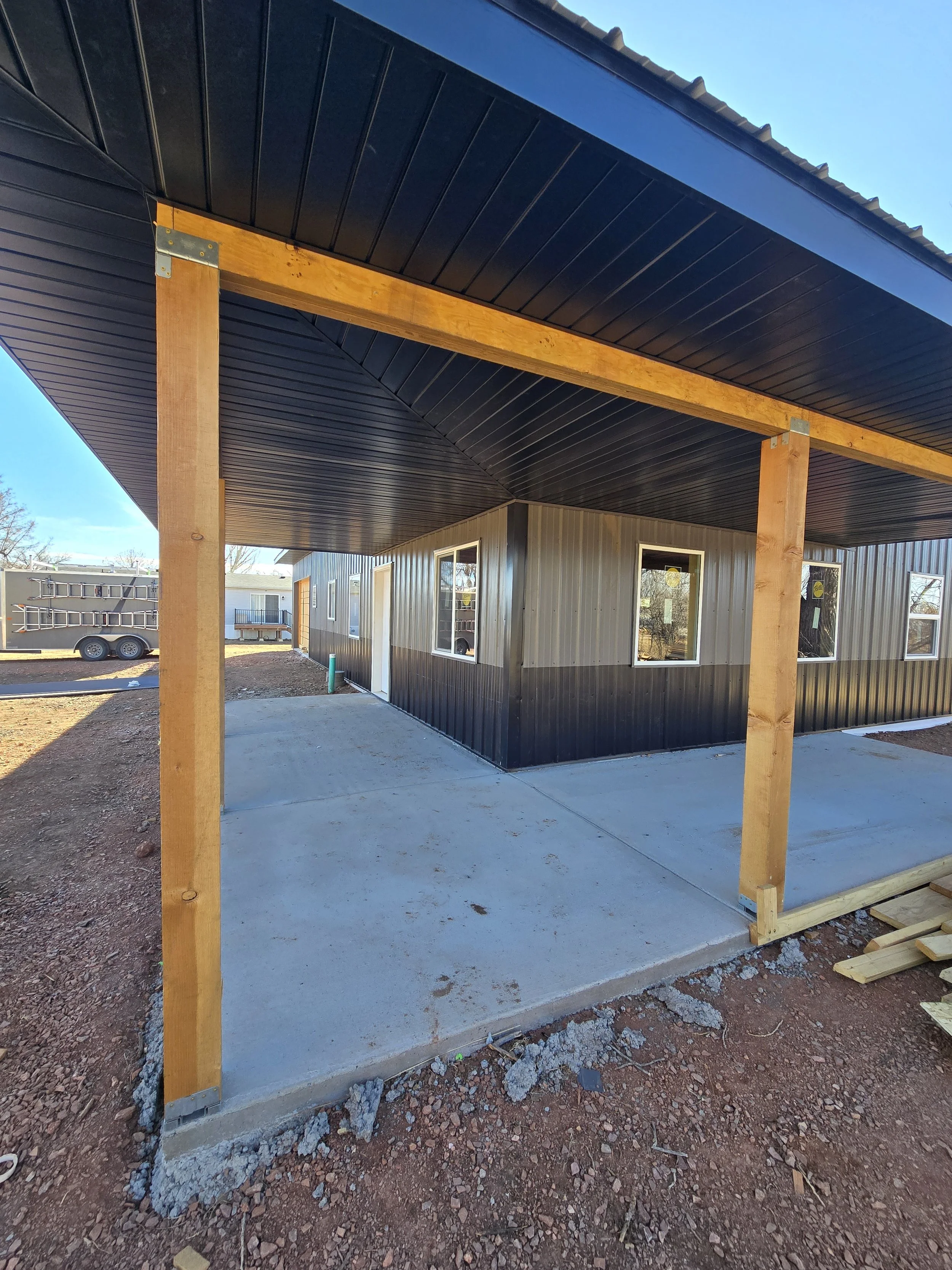Construction site showing a concrete patio with an unfinished wooden structure supporting a black metal roof overhang, next to a building with gray siding and white-framed windows.