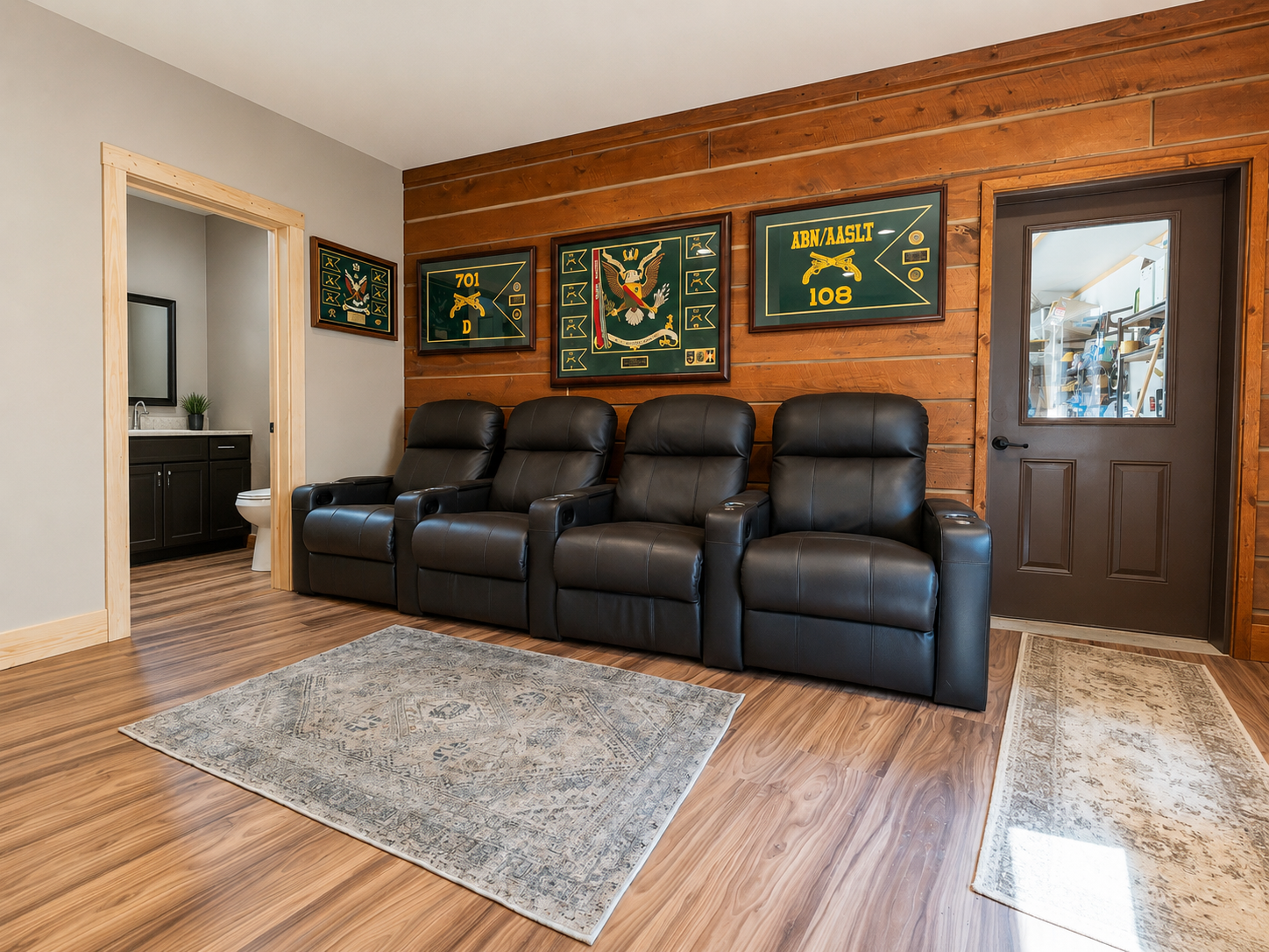 Room with four black leather recliners arranged in a row, behind a wooden wall with military-themed artwork, and a door leading to a storage room. A gray and beige patterned rug on a wooden floor.