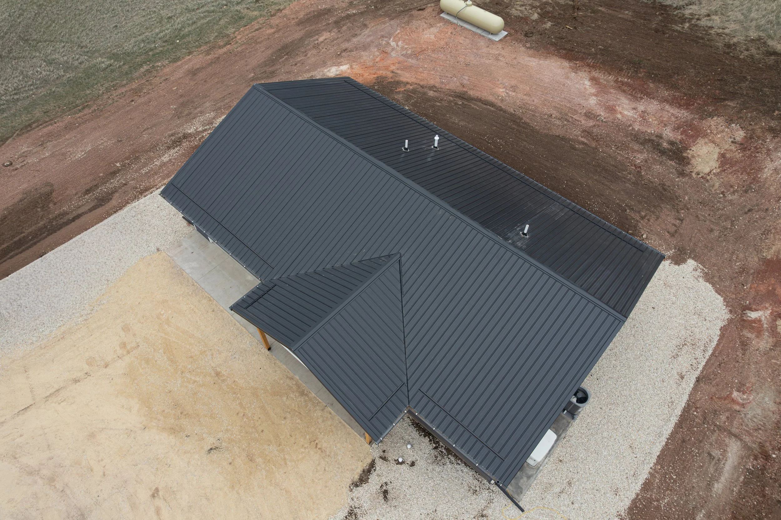 Aerial view of a newly built house with a dark metal roof, on a construction site with dirt and gravel ground, and some industrial tanks nearby.