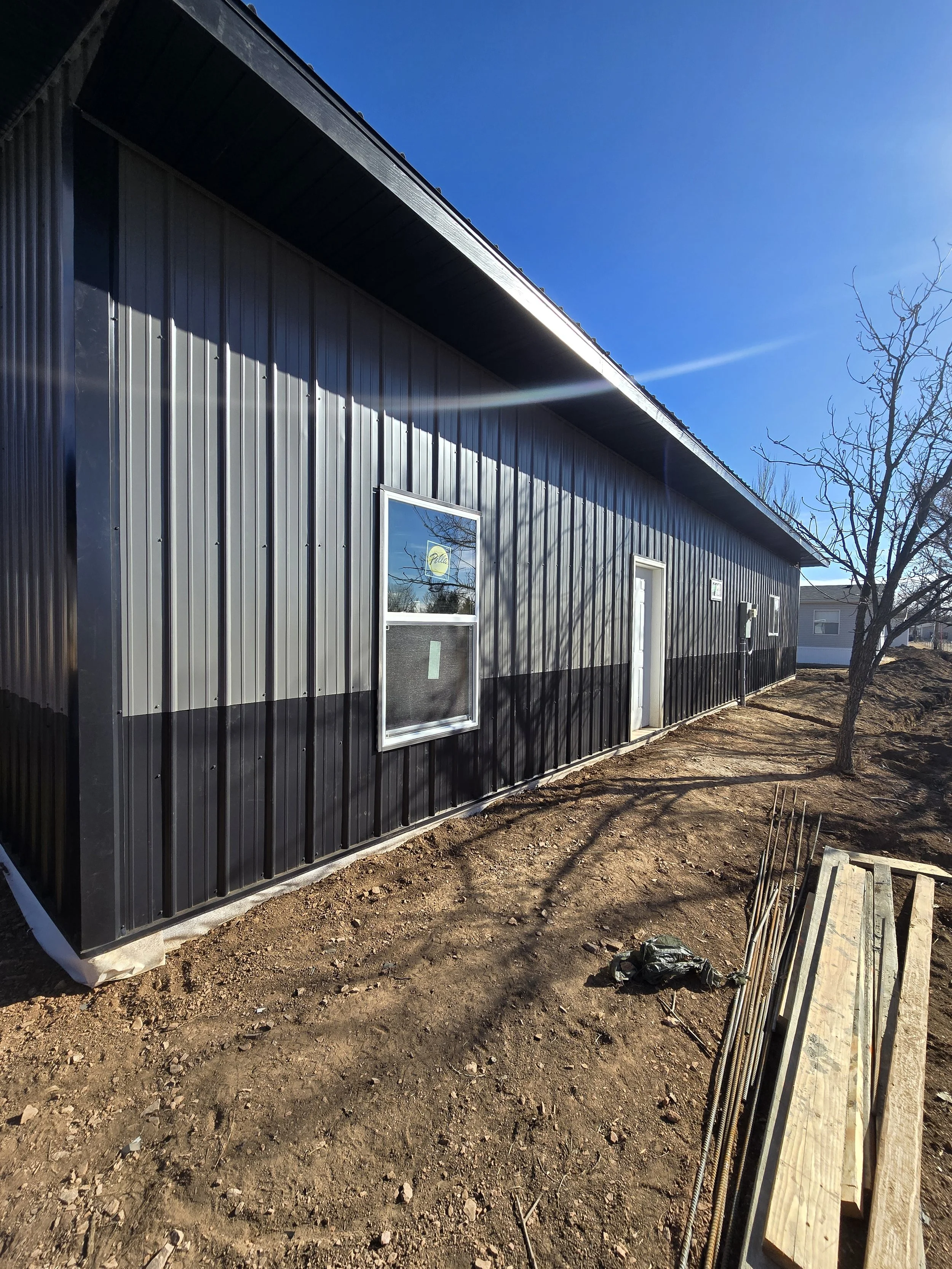 A black metal building with three windows and a door, partially under construction, with exposed soil and some construction materials nearby. The sky is clear and blue, and the building is casting shadows.